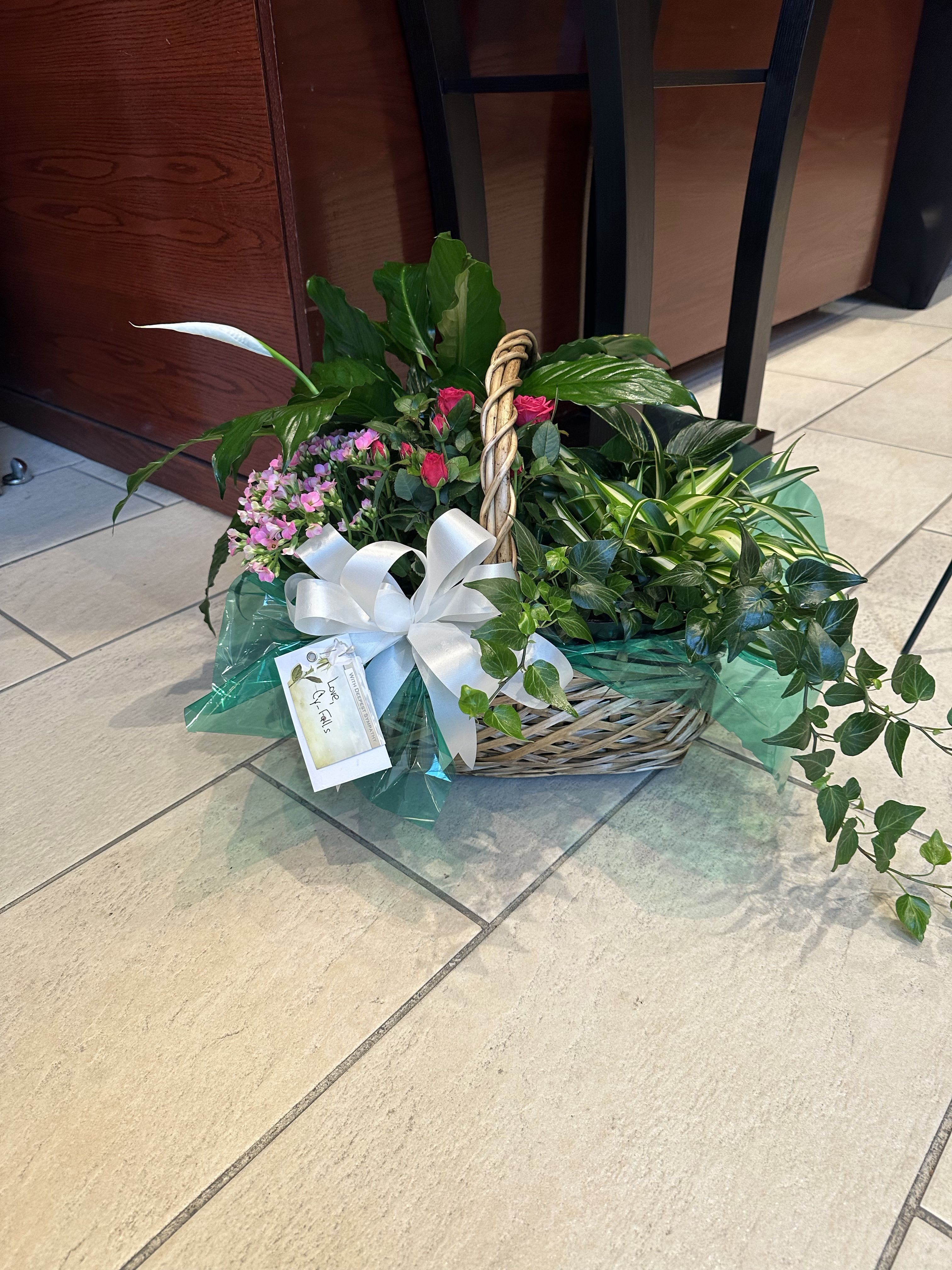 Basket garden of assorted green plants with pink blooms, a peace lily, and a white bow.