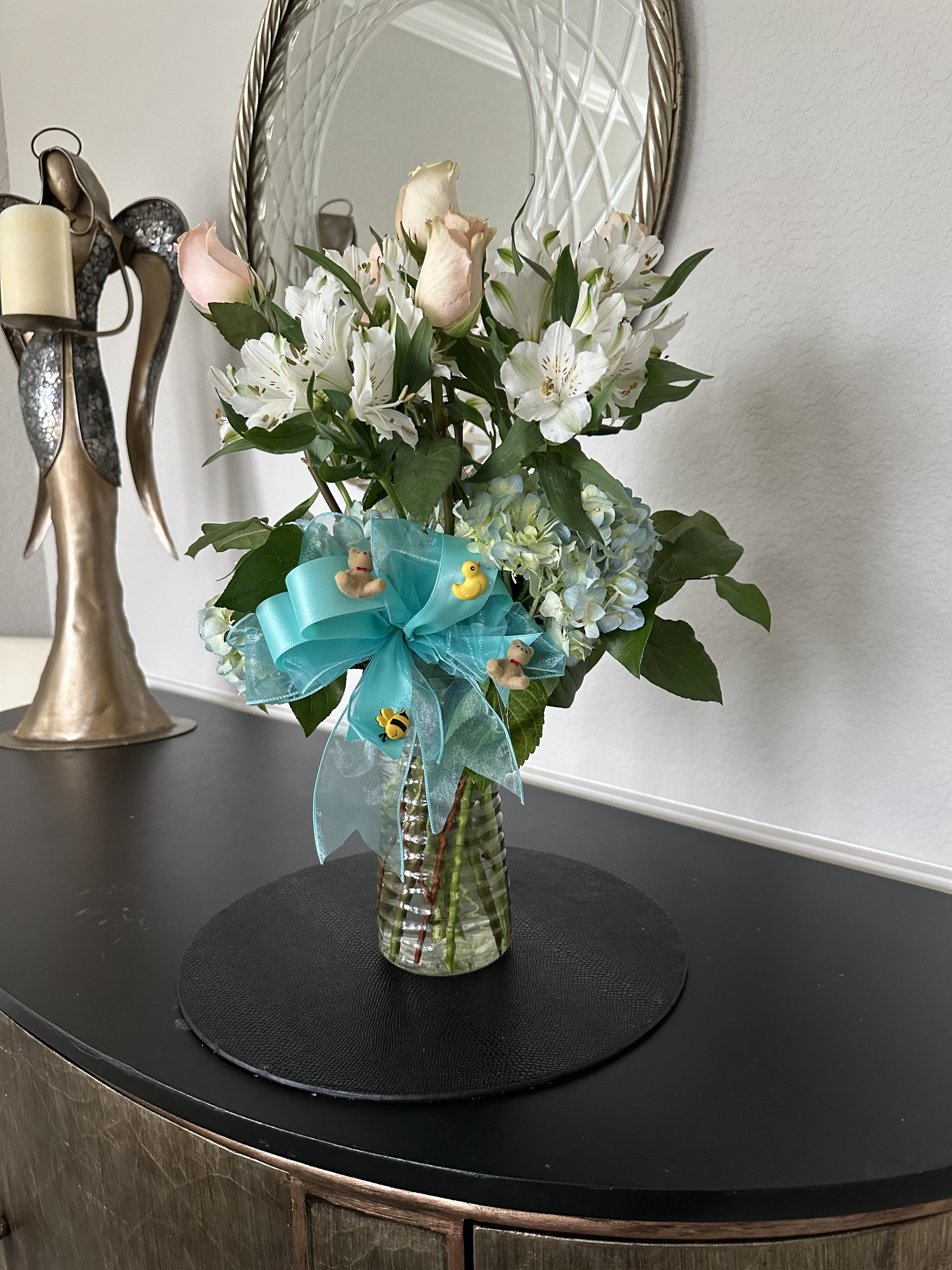 Vase arrangement of white alstroemeria, pale pink roses, and blue hydrangea with a teal bow and small baby-themed decorations.