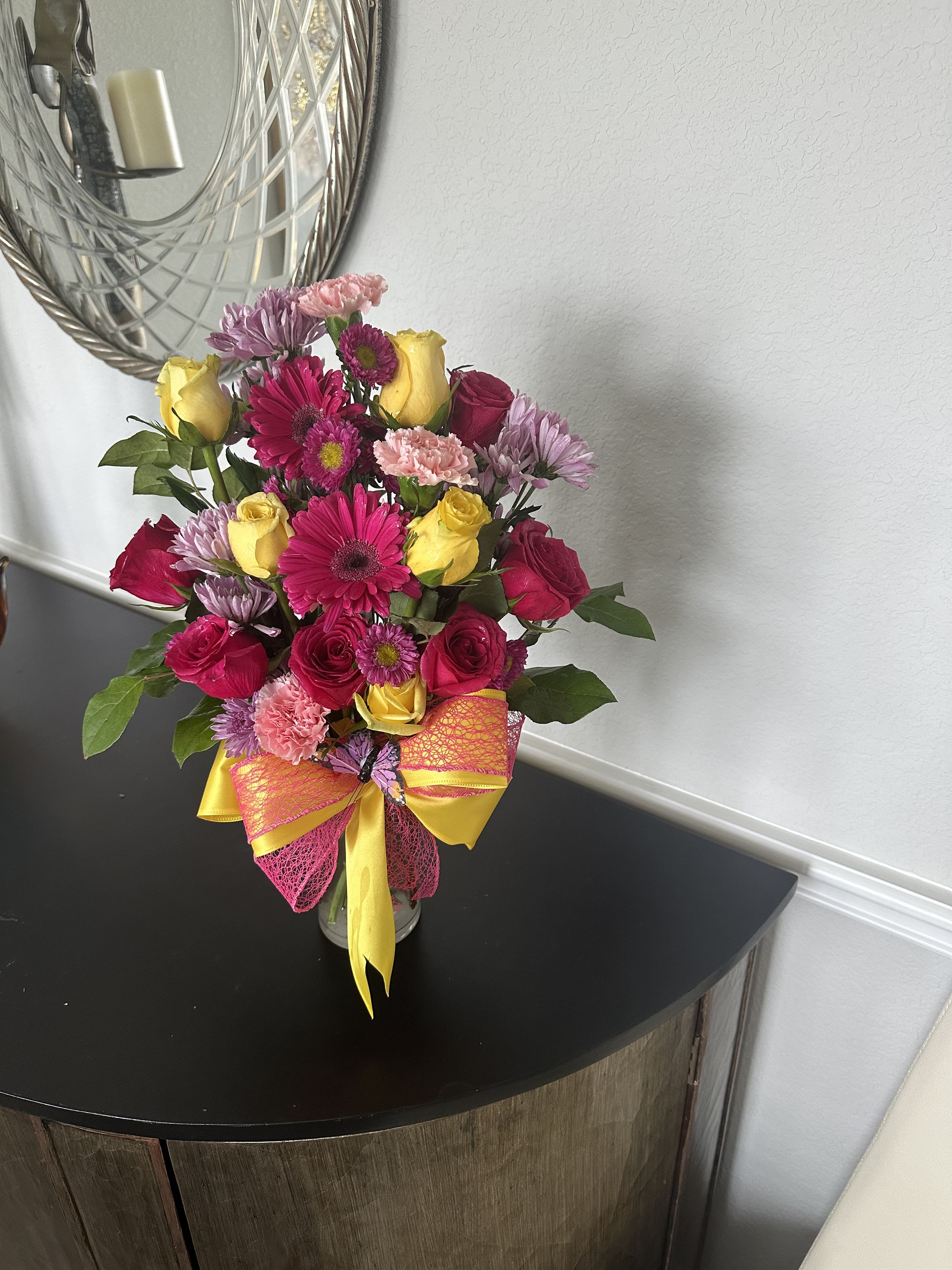 Bright mixed bouquet of pink roses, gerbera daisies, and yellow roses in a clear vase with a yellow and pink bow.