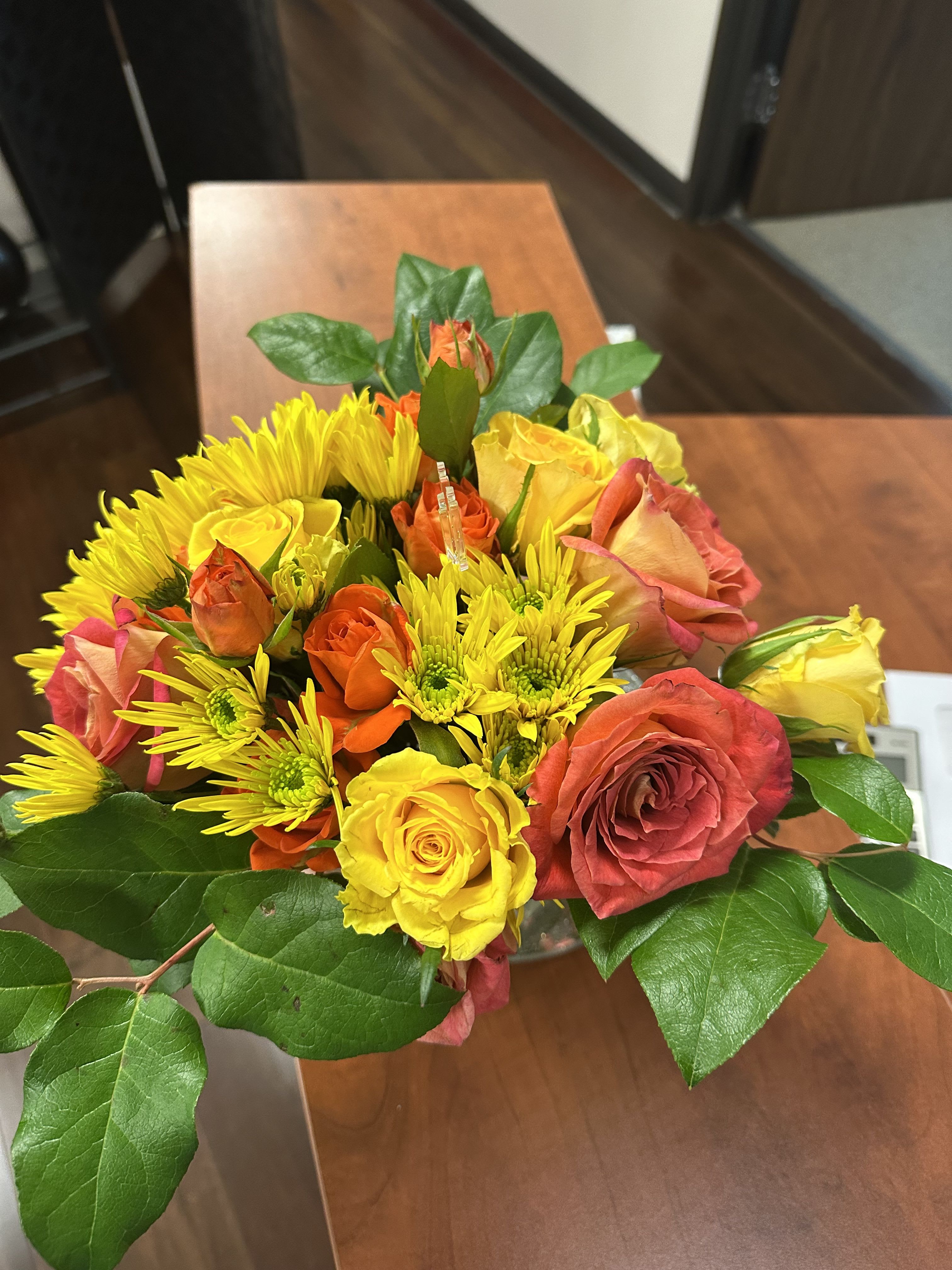 Low arrangement of yellow and orange roses and yellow mums in a clear glass vase on a wooden table.