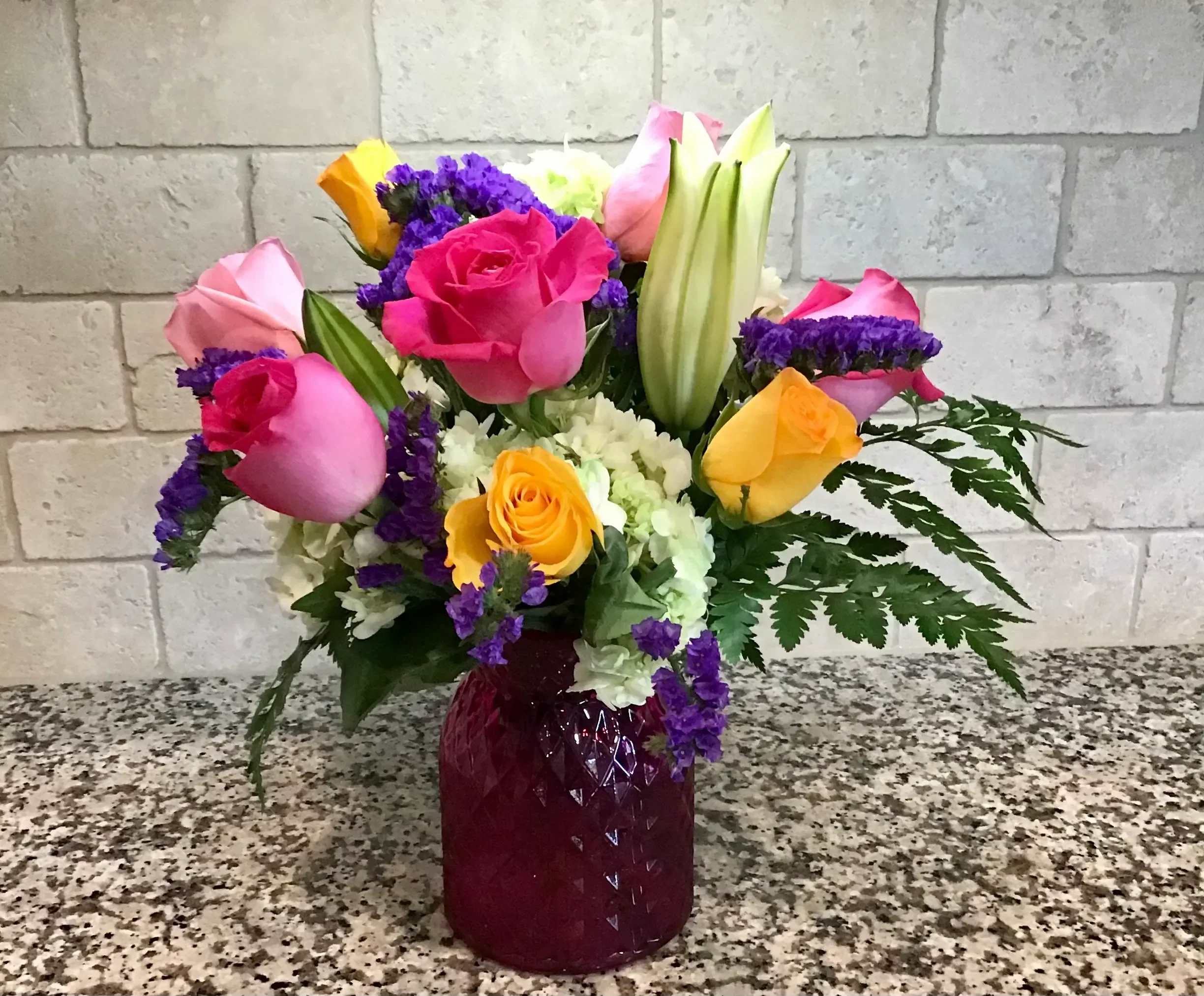Mixed bouquet of pink and yellow roses, lilies and white blooms in a red glass vase on a speckled counter.