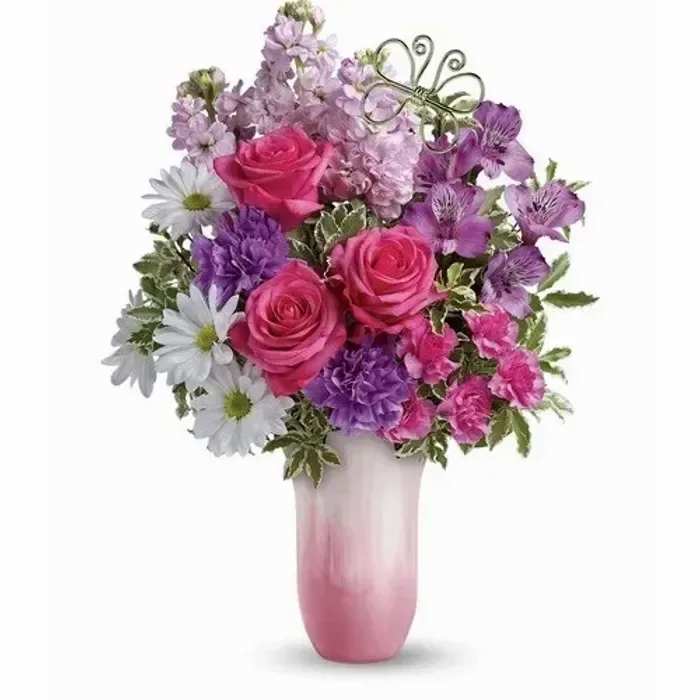 Pink roses, purple blooms, and white daisies in a pink ombré vase with a metal butterfly accent