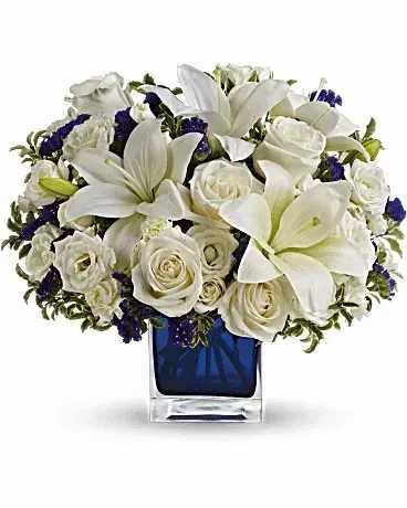 Low arrangement of white lilies and roses with purple accents in a blue cube glass vase
