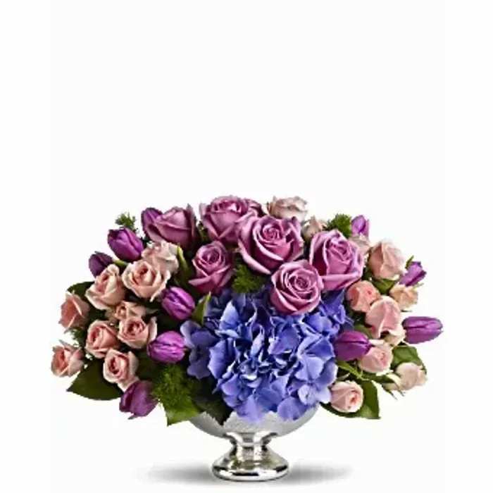 Low centerpiece of purple roses, pink spray roses, tulips, and blue hydrangeas in a silver pedestal bowl