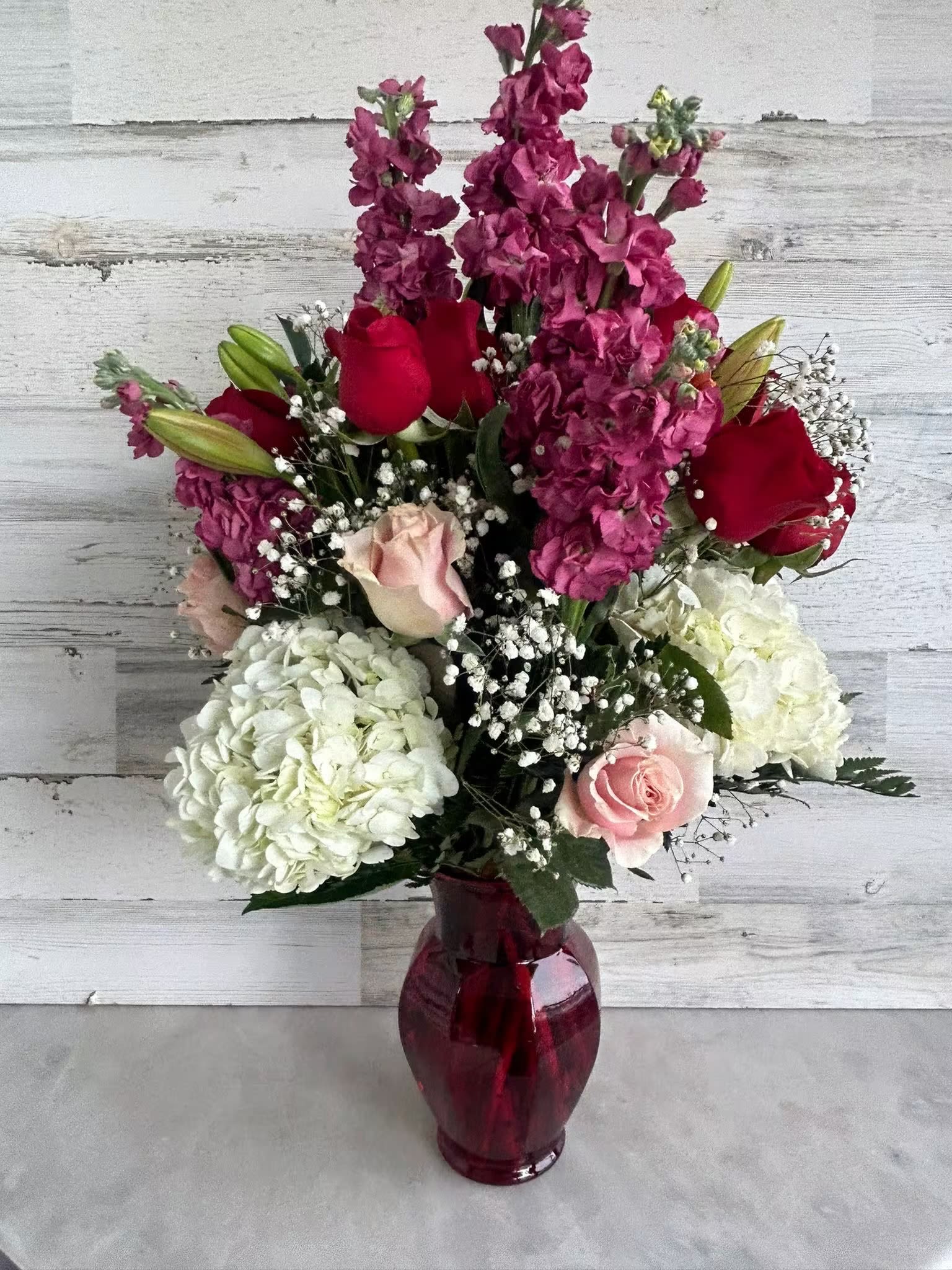 Tall arrangement of pink stock, red and pink roses, and white hydrangeas in a red glass vase