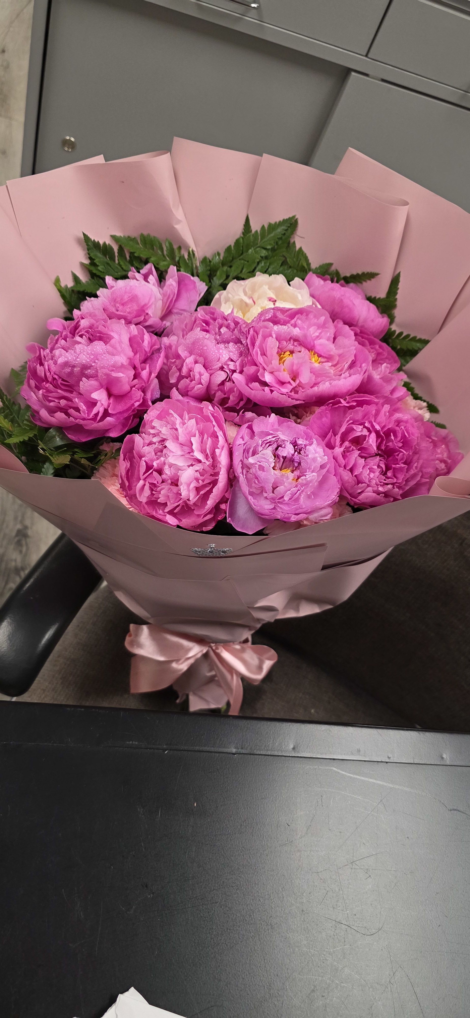 Ramo de peonias  - Lush, romantic pink peonies take center stage in this birthday bouquet, their ruffled petals forming a full, cloud-like cluster that immediately reads as celebratory and feminine. A single pale cream peony near the top adds a soft highlight that catches the eye-perfect for an anniversary, Mother's Day, or any moment you want the person you're thinking of to feel cherished.  Dark green foliage peeks out around the blooms, giving the bouquet fresh contrast against the blush wrapping paper and matching satin ribbon for a polished finish. The arrangement is hand-tied and presented in decorative paper that keeps the blooms secure while looking elegant at first glance.  Send this bouquet to mark a milestone, say "thank you," or simply make someone feel truly seen; it's the kind of gift that reads as thoughtful and joyful the moment it's handed over. Care: keep peonies in fresh water, trim stems at an angle, and place away from direct heat to prolong bloom life.