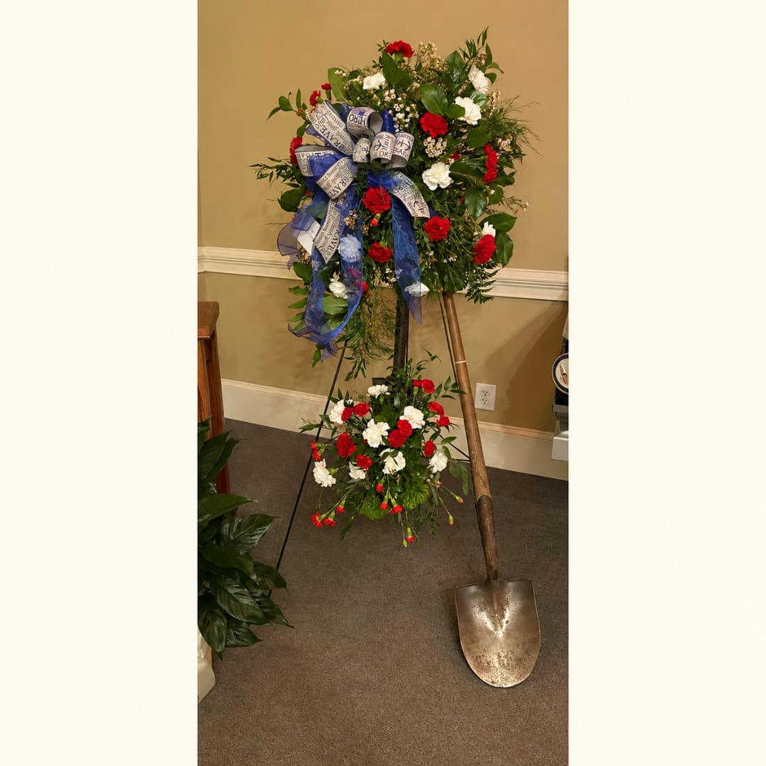 Standing floral spray with red and white carnations and a blue ribbon on a shovel