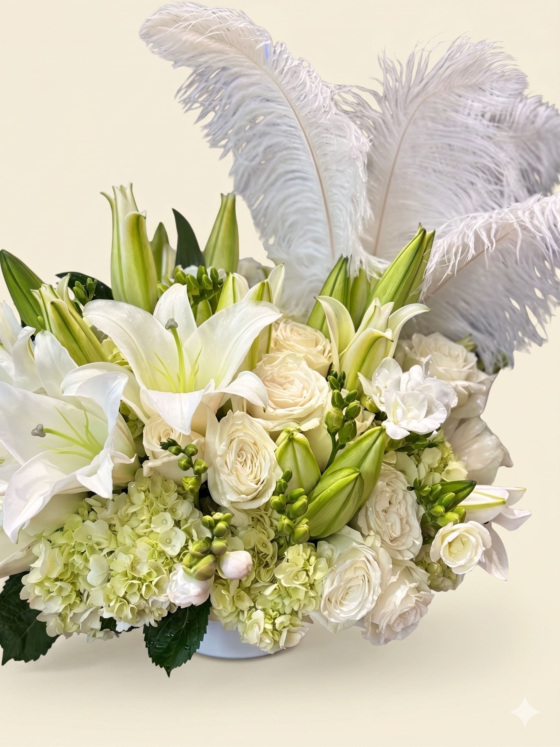 BP78 - Feathered Grace Arrangement - This elegant all-white arrangement features graceful lilies, lush roses, and soft hydrangeas, accented with delicate white feathers. Its airy beauty and timeless charm create a sense of peace and sophistication.  White feathers are optional. 