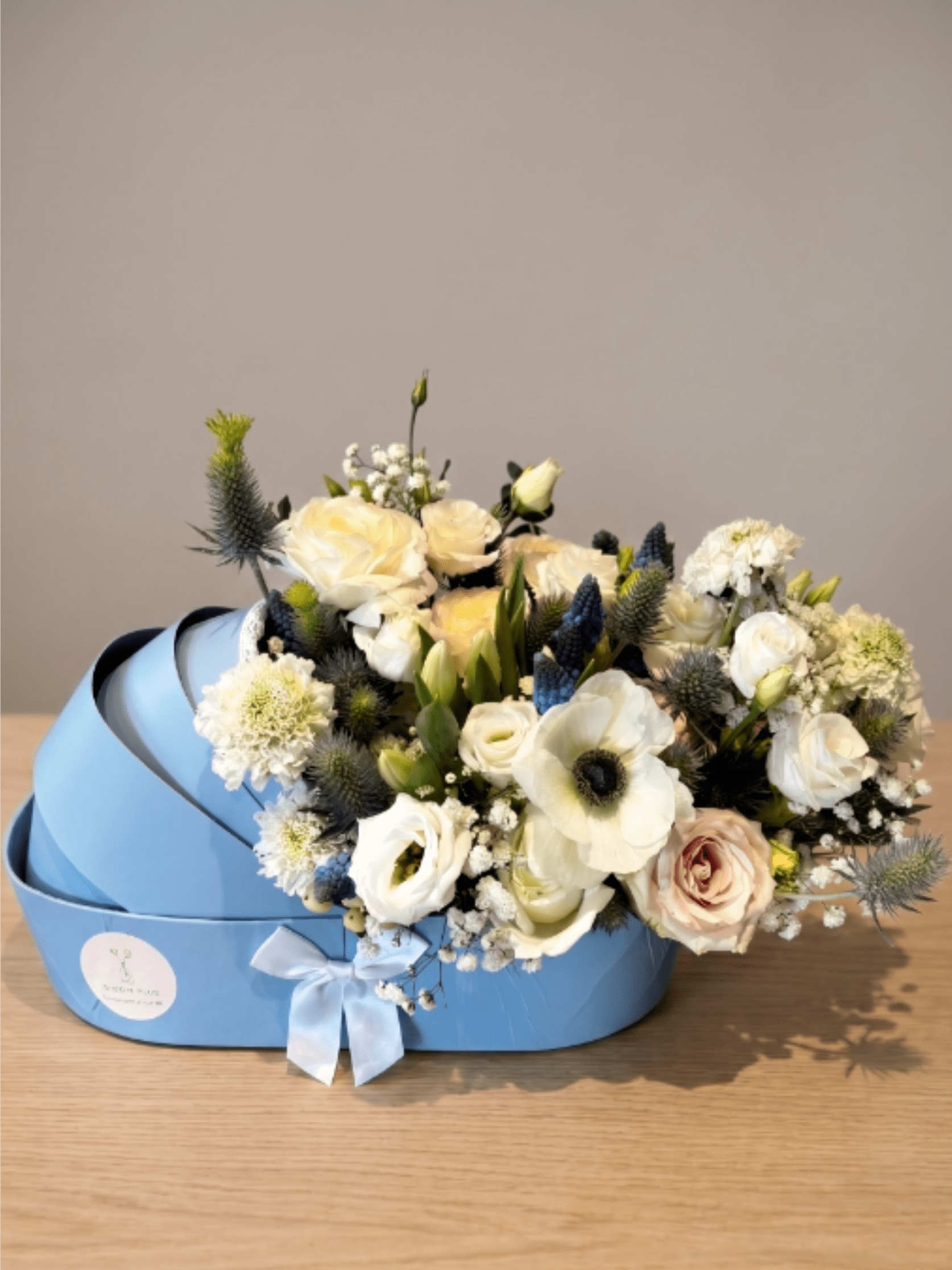 BP110 - Little Blue Carriage - This sweet, whimsical arrangement gathers white anemones, creamy roses, and textured white scabiosa, perfectly accented by striking blue thistle, delicate muscari, and airy baby's breath in a charming, light blue baby carriage box.  Seasonality: Anemones: Peak availability in late Winter through Spring. Muscari: Early Spring bloom. Scabiosa: Late Spring through Summer. Blue Thistle (Eryngium): Available year-round. Roses &amp; Baby's Breath: Available year-round.