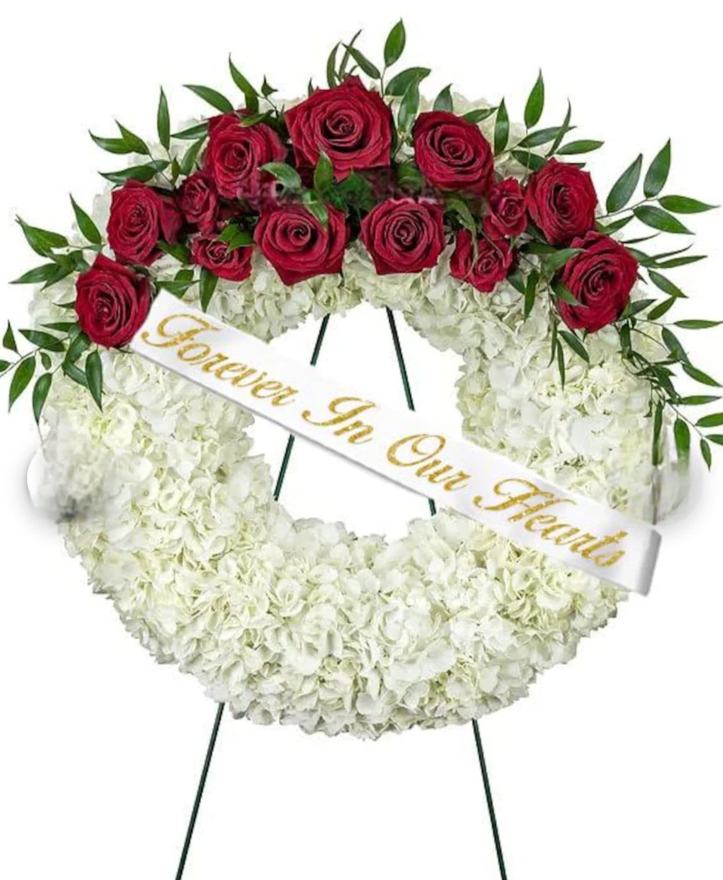 Infinity Love - A circular standing wreath of white crisantemos and red roses designed for sympathy and funeral services, this arrangement offers a classic, dignified tribute that reads as both timeless and comforting. The white base gives a cloud-like, textured softness while the bold band of deep red roses across the top arc creates a focused, heartfelt accent-an arrangement that lets the person you're honoring feel remembered and deeply cherished.  The wreath is densely covered in small white blooms with a row of rich red roses interspersed with slender vertical accents to add height and movement. A white memorial ribbon with gold script reading "Forever In Our Hearts" is draped diagonally across the face, and the arrangement is presented on a slim green metal easel for formal display. Dimensions and exact stem counts are available on the product page; the sturdy easel and secure wiring make it suitable for services, memorial gatherings, or placement at a graveside.  Appropriate for funeral services, commemorative events, or as a respectful centerpiece at a memorial, this wreath communicates sympathy and lasting affection for the person you're honoring. For delivery and placement guidance, or to request ribbon wording alternatives, contact us at checkout so we can ensure the tribute arrives exactly as you intend.