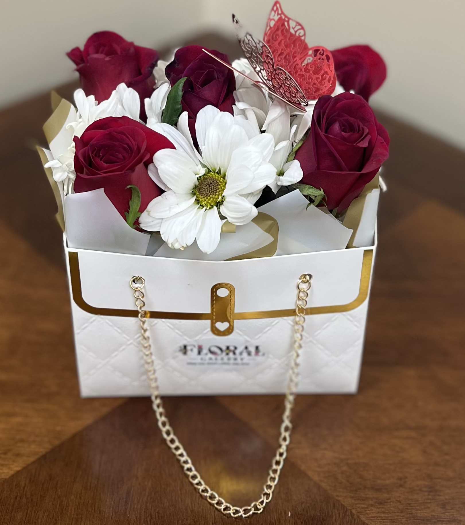 Whispers of Love by Floral Gallery - A luxurious blend of deep red roses and pure white daisies, artfully arranged in a white handbag box with gold accents and a delicate butterfly touch. This elegant design speaks of passion and grace, perfect for expressing love, admiration, or heartfelt appreciation with timeless charm.
