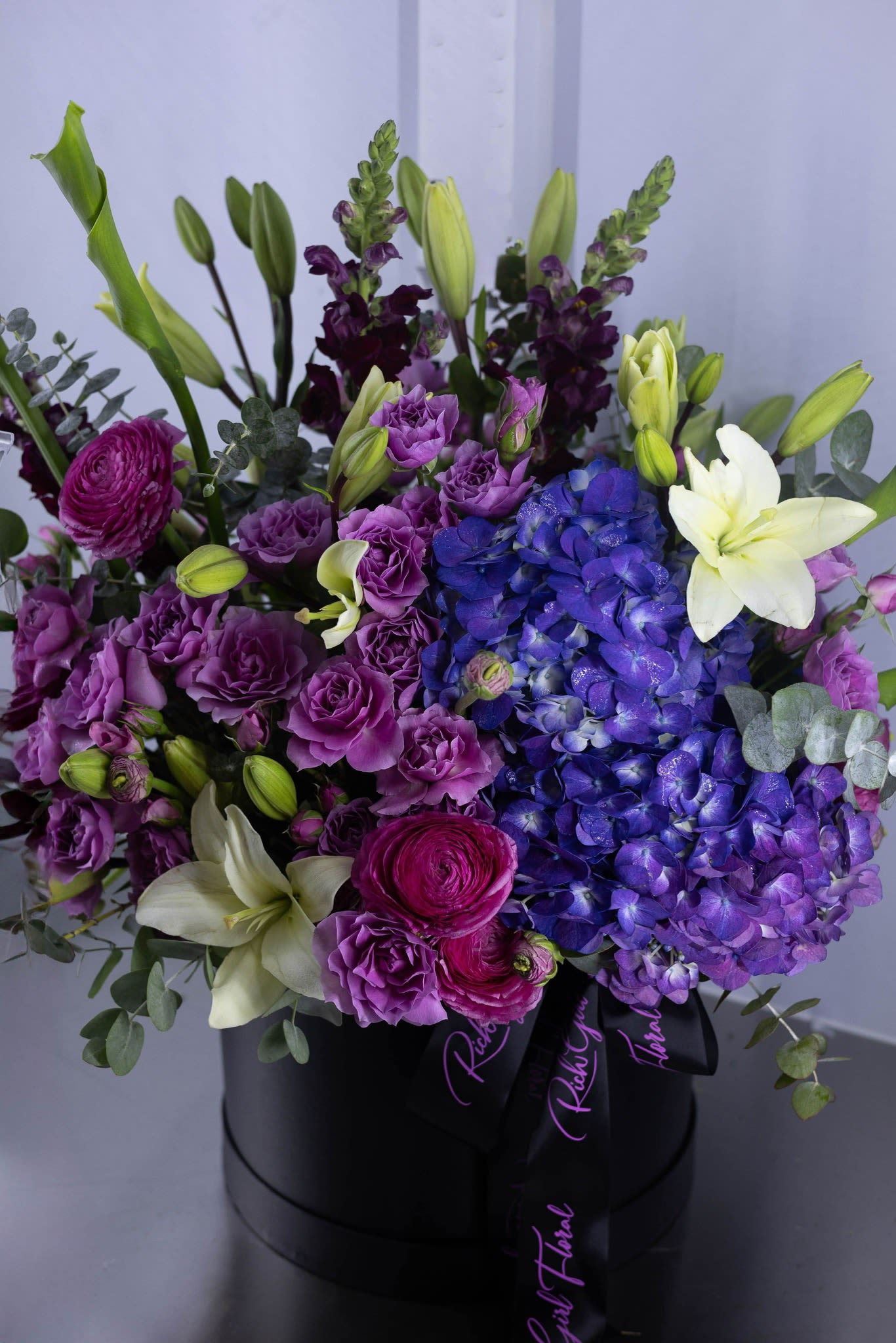 Purple and blue floral arrangement with white lilies in a black hat box