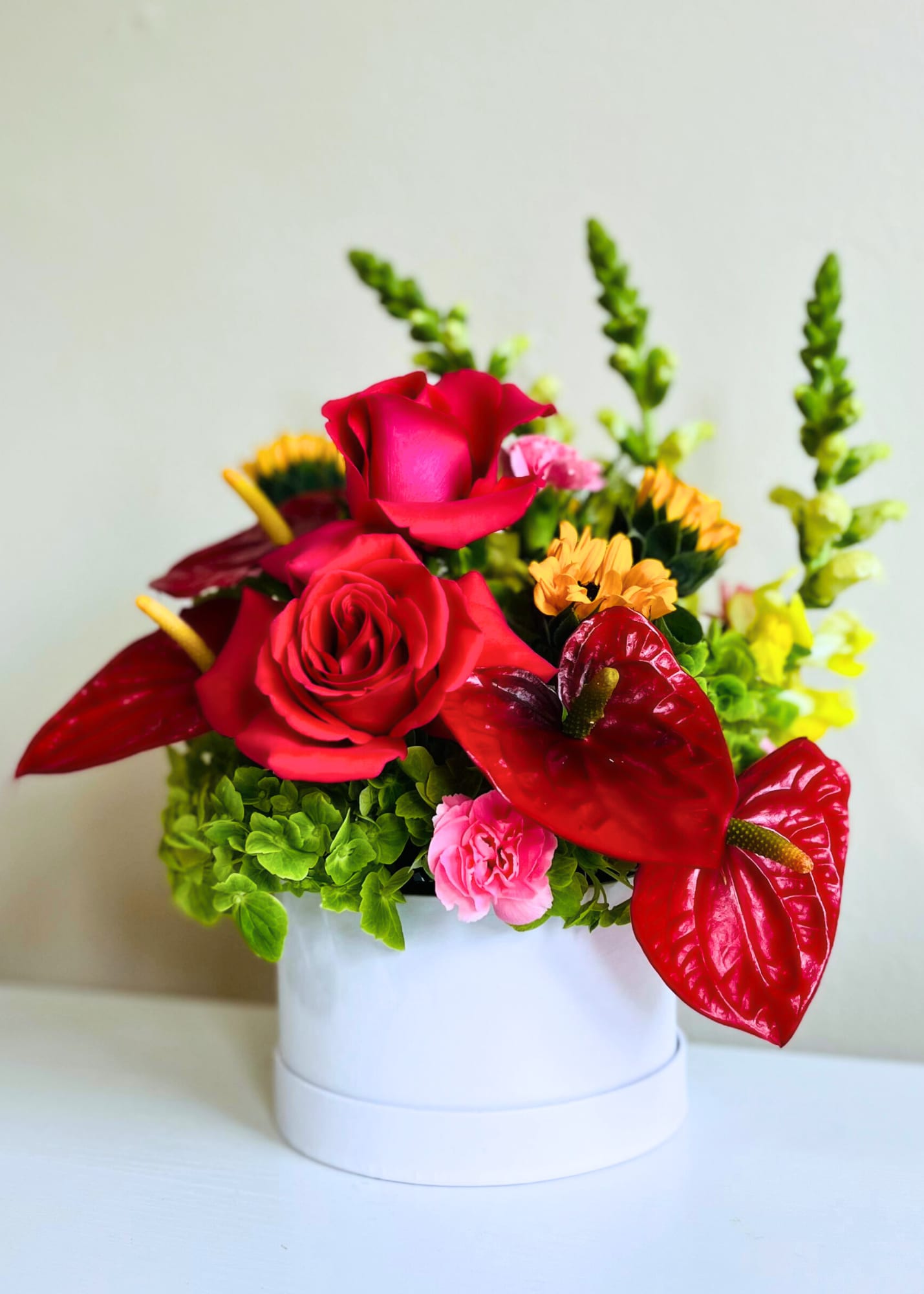 Compact arrangement of red roses and anthuriums with mixed bright blooms in a white round container