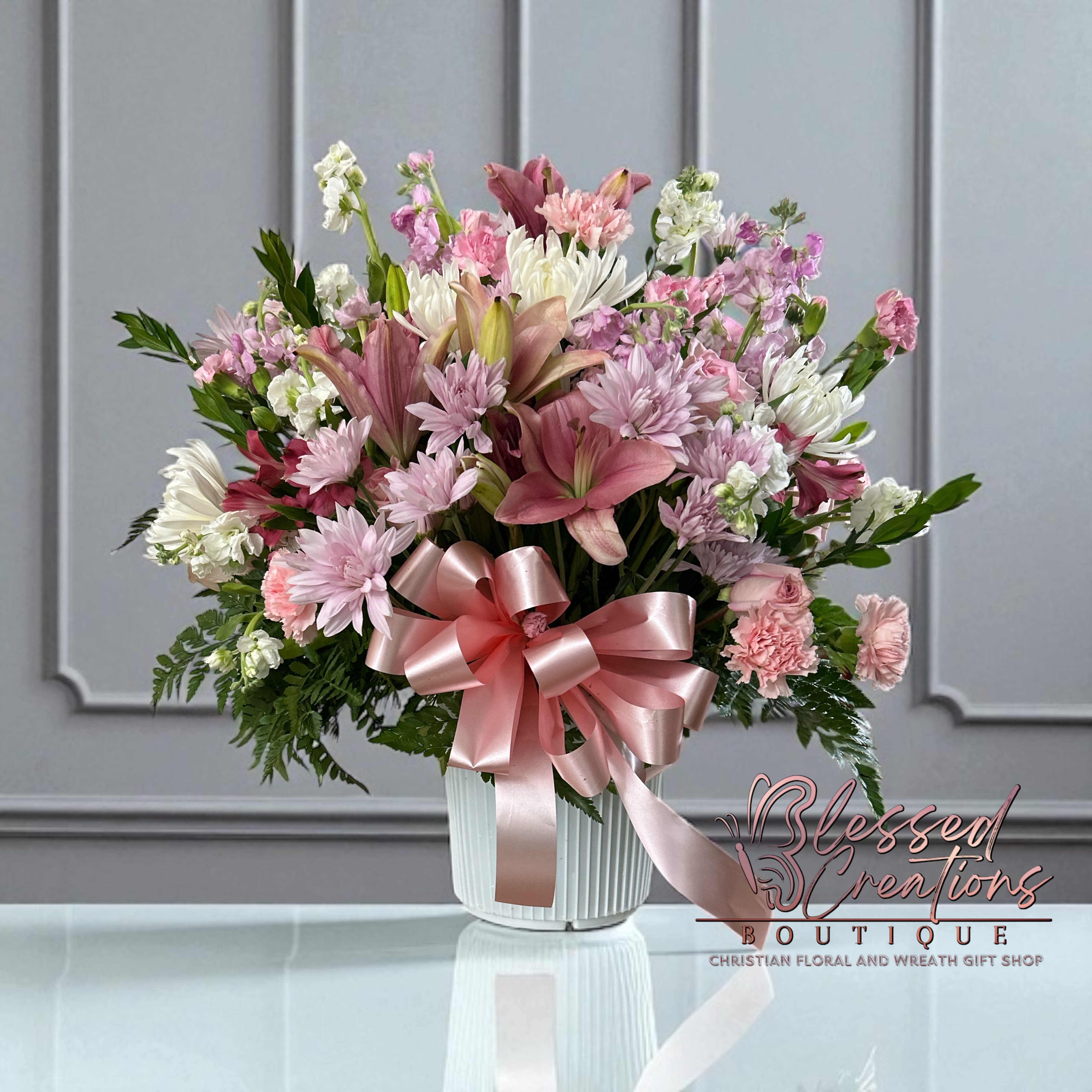 Graceful Pink Tribute  - The Graceful Pink Tribute is a soft and elegant display of love and comfort. Featuring pink and lilies, carnations, daisies, stock, and white lilac accent flowers, all designed with lush greenery and finished with a beautiful pink satin bow. Presented in a classic white container, this arrangement is a touching expression for sympathy, memorial services, or to simply share comfort and hope.