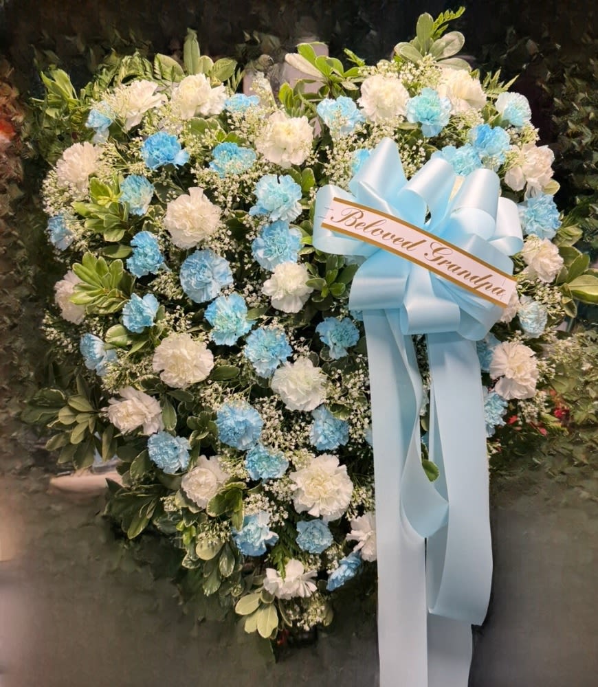 Full of Blooms - "Offer a heartfelt and serene tribute with our 'Beloved' Heart-Shaped Sympathy Spray. This exquisite floral arrangement, shaped to symbolize enduring love and cherished memories, features a harmonious blend of pristine white and calming blue blooms, accented with lush greenery and delicate baby's breath. A prominent light blue ribbon proudly displays a message,  making this a profoundly personal and beautiful expression of remembrance. Perfect for honoring a life well-lived and conveying deep condolences during a difficult time."