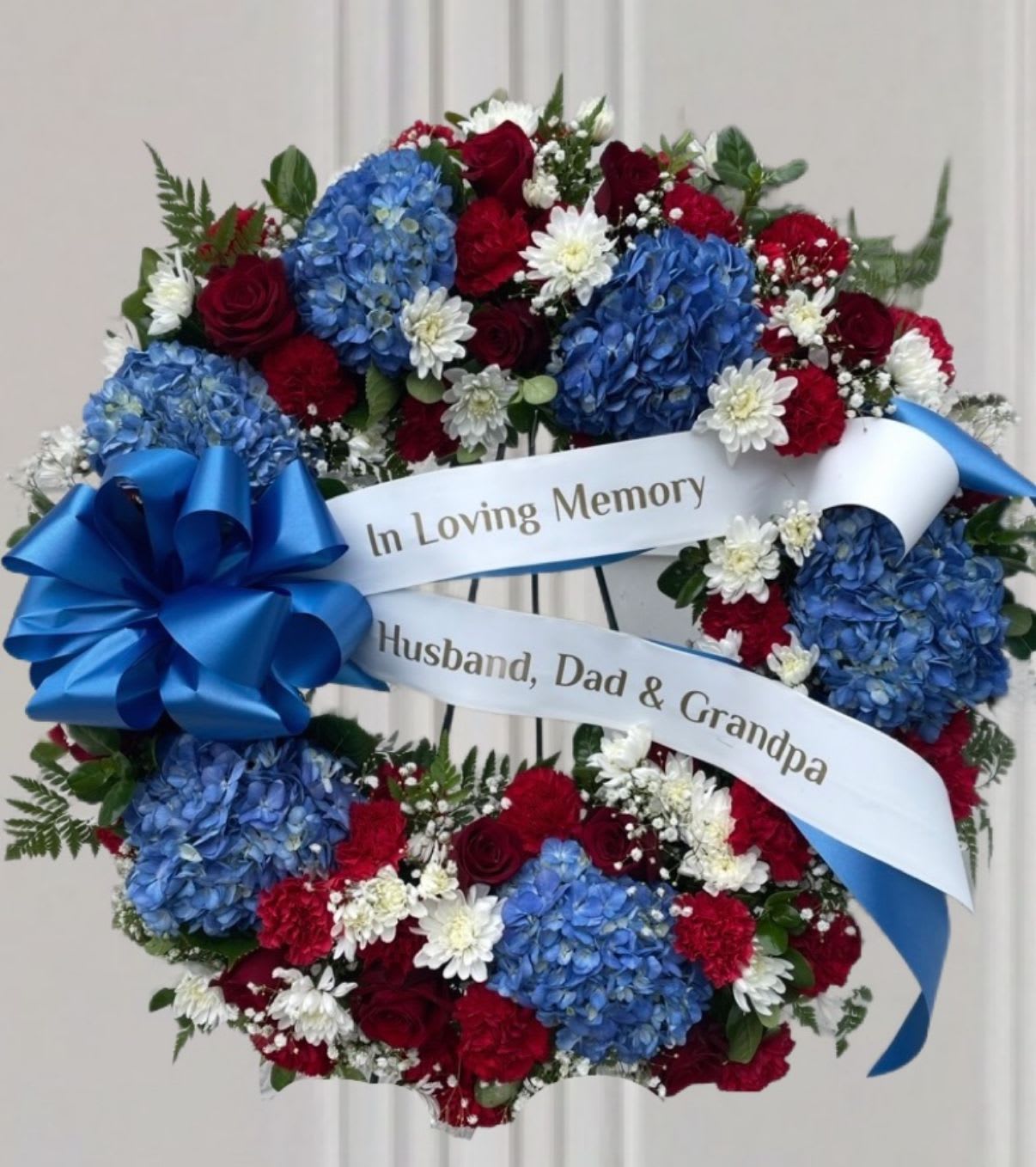 American Tribute - "Honor the memory with our exquisite Patriotic Remembrance Wreath. This stunning floral tribute features a lush arrangement of rich red roses and carnations, serene white chrysanthemums, and striking blue hydrangeas, creating a powerful display of national pride and enduring love. The circular design symbolizes eternal life and the unbroken bond of remembrance, providing comfort and solace during a time of loss. This wreath serves as a deeply moving and personal expression of honor and affection for a life beautifully lived."