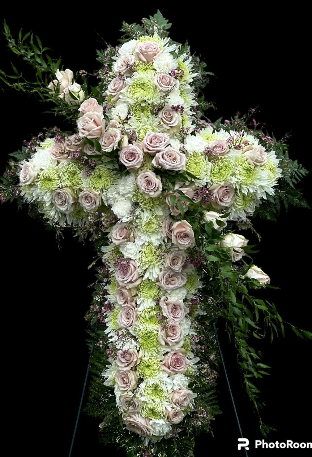 Eternal Grace Cross - In times of remembrance and reflection, our "Eternal Grace" Floral Cross offers a profound and elegant tribute. This exquisite standing spray, meticulously crafted in the sacred shape of a cross, serves as a powerful symbol of faith, hope, and everlasting peace. Designed with a harmonious blend of soft hues, this arrangement features an abundant base of serene white and gentle green chrysanthemums, creating a lush, comforting foundation. Delicately woven throughout are graceful blush-pink roses, adding a touch of tender love and profound respect. Lush green foliage frames the arrangement, lending a natural beauty and depth to this heartfelt expression.