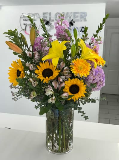 Bright Days Ahead - Like a breath of fresh air from a blooming summer field, this tall and cheerful bouquet features vibrant sunflowers, bright lilies, soft purple stock, and delicate alstroemeria. Arranged in a clear glass vase, it brings warmth, light, and joy to any room or celebration. (If some flowers and/or the vase are not available there will be minor substitutions.) 