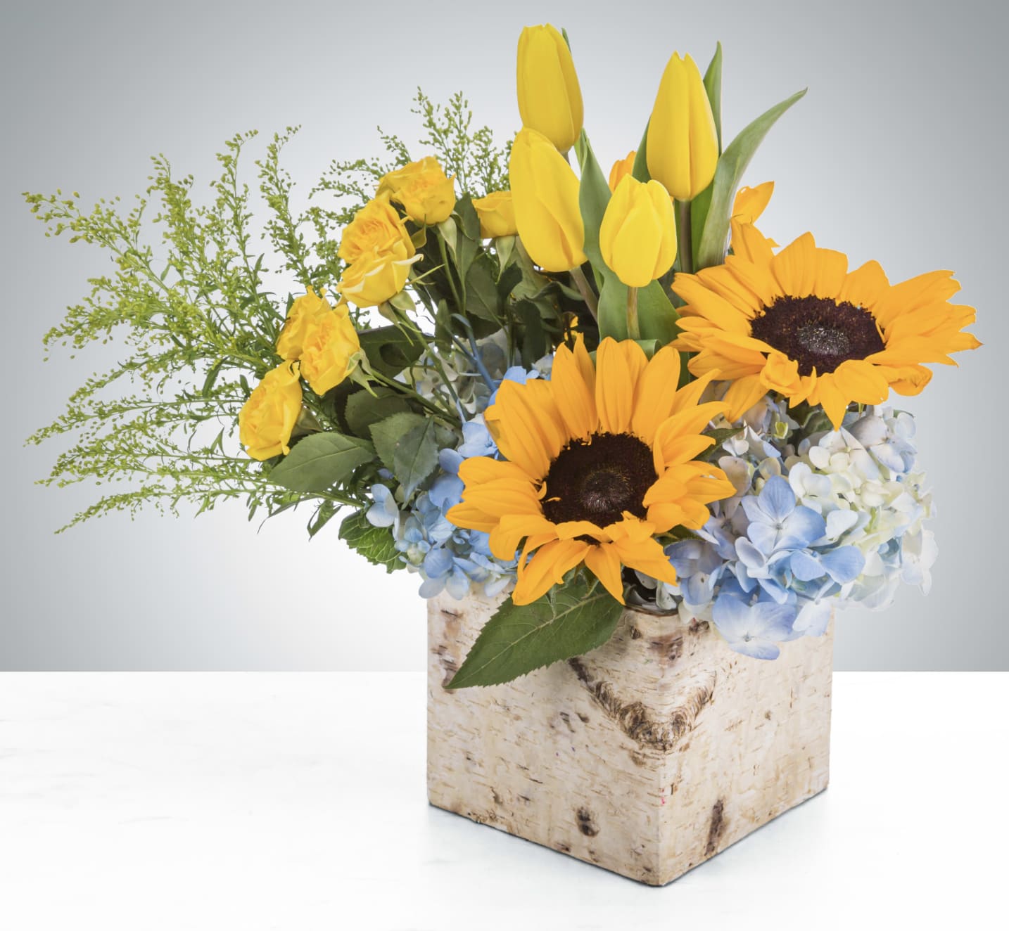 Yellow tulips, roses, sunflowers, and blue hydrangeas in a birch-style cube vase