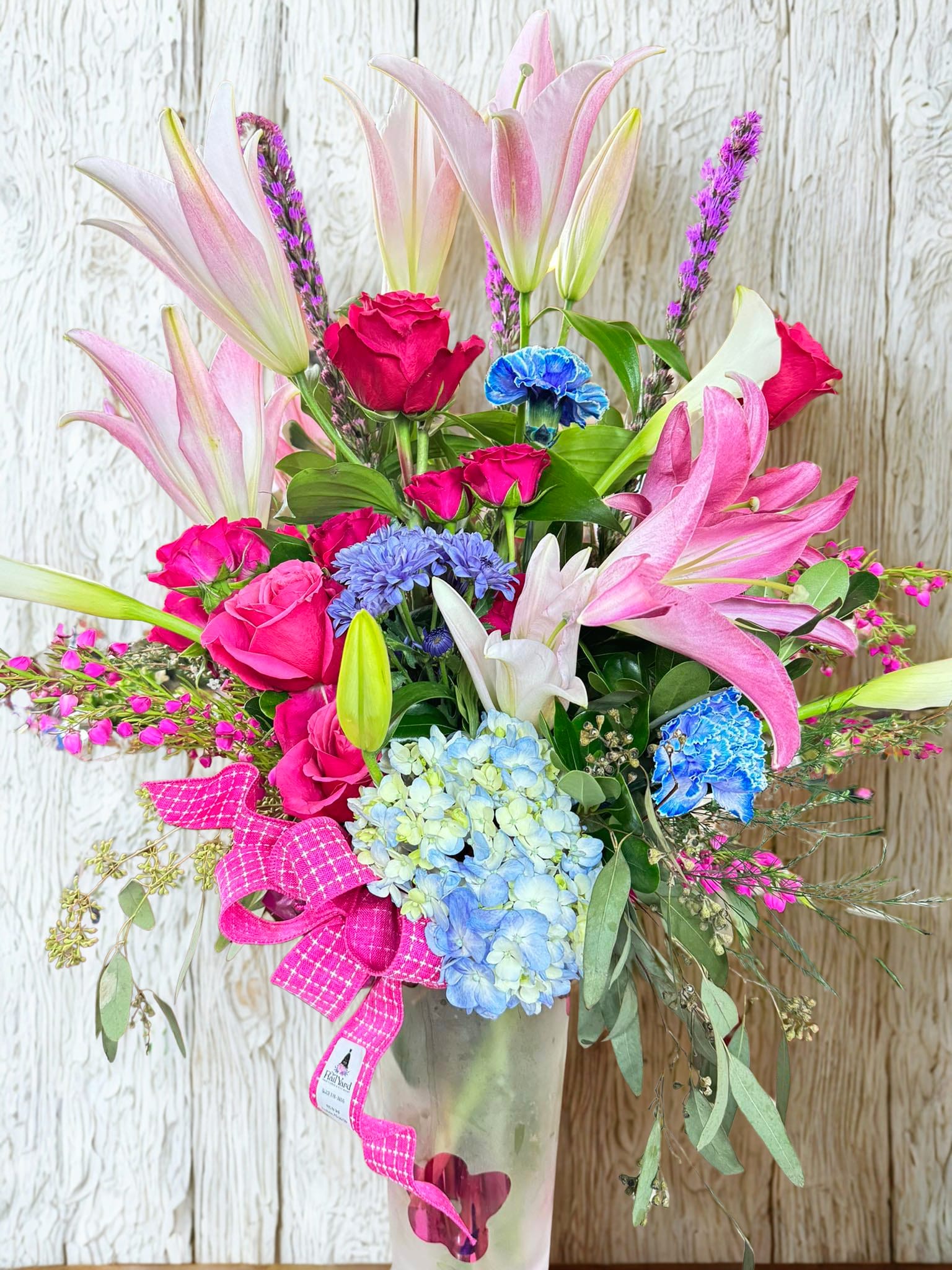 Pink lilies, roses, and blue hydrangeas in a glass vase with a pink ribbon