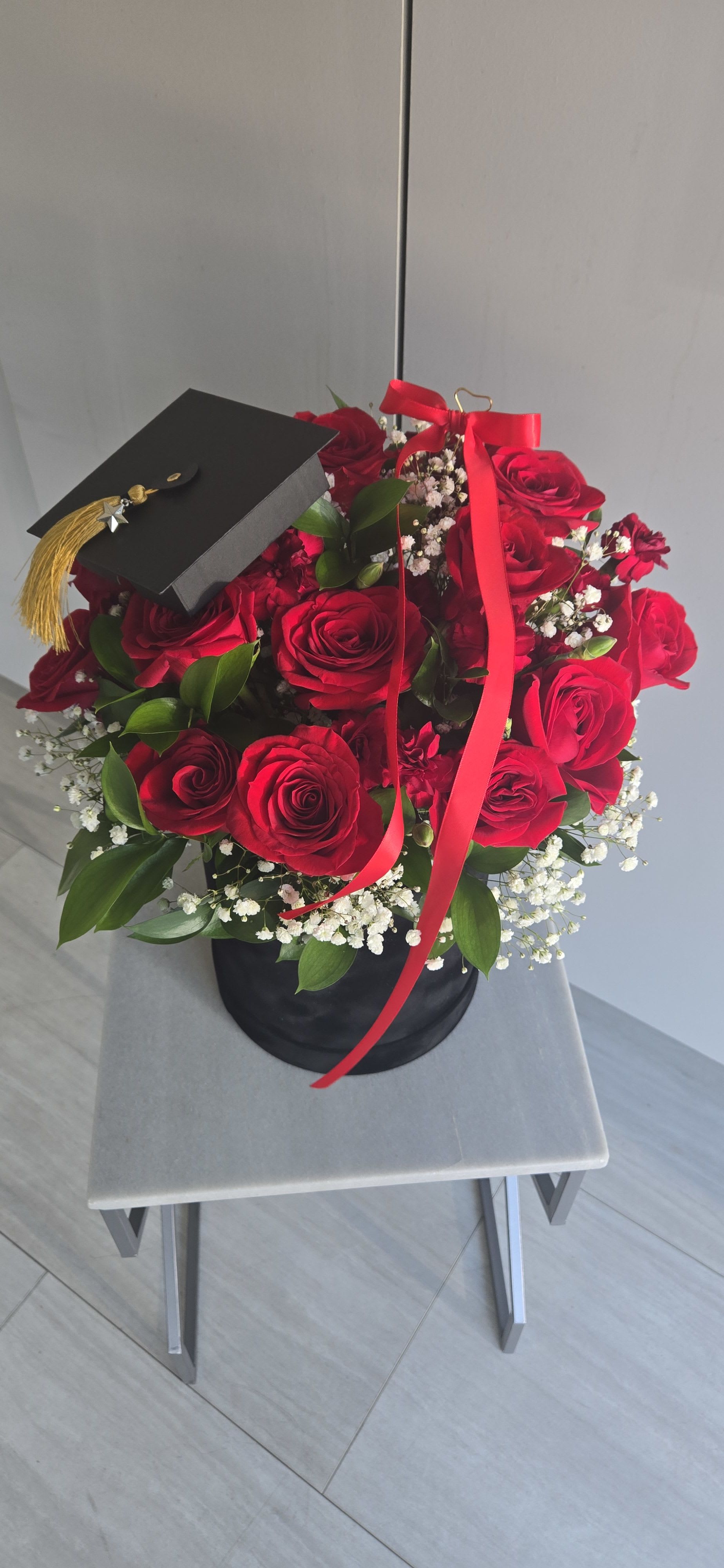 Graduation  - "Celebrate success in style with this elegant floral arrangement. Featuring lush red roses, delicate baby’s breath, and fresh greenery, all beautifully presented in a sleek black hat box. Complete with a mini graduation cap and congratulatory card, it’s the perfect gift to honor a proud graduate’s big milestone." 