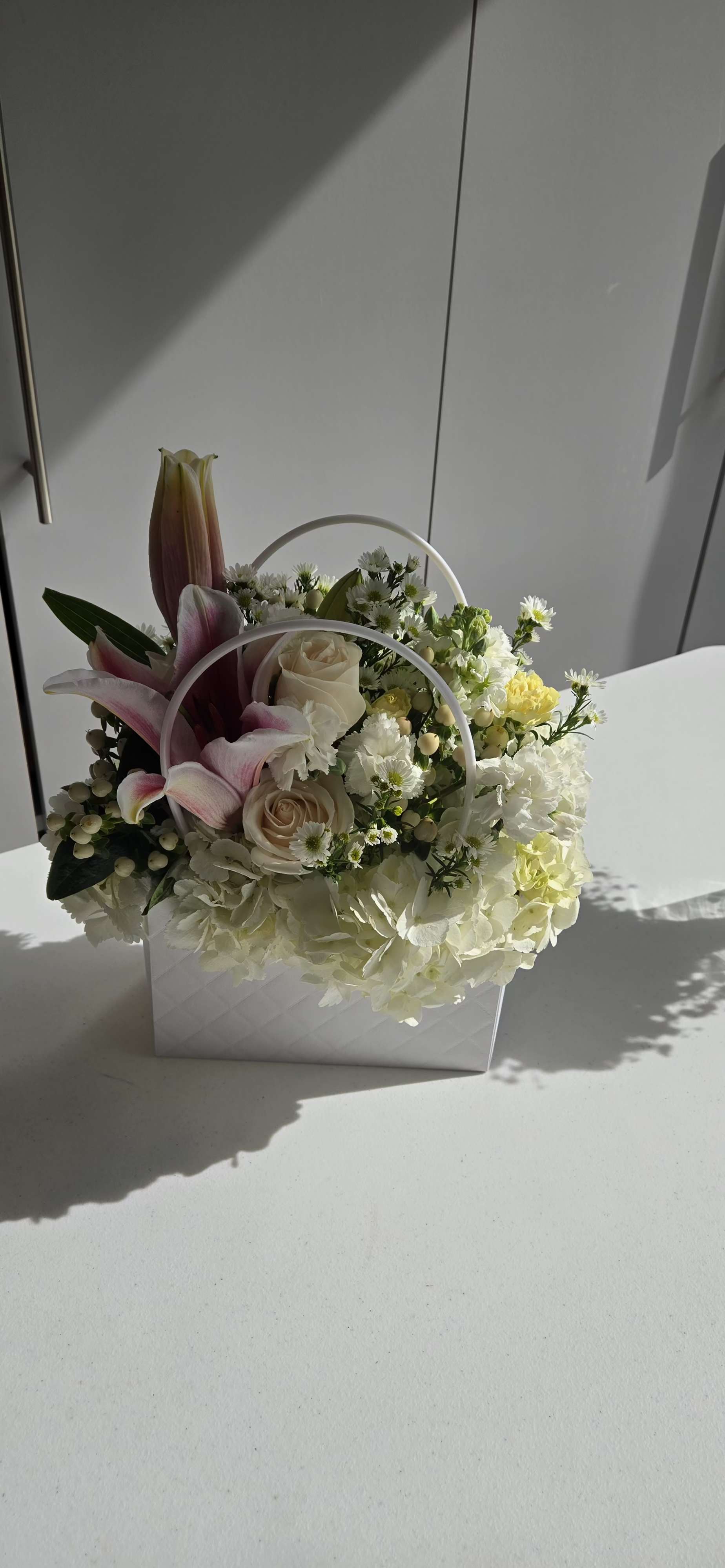 Floral bag  - This beautiful floral arrangement features a soft and elegant palette of cream, white, and blush tones. The mix includes pale pink lilies, cream roses, delicate white daisies, hydrangeas, and pops of yellow blooms, all thoughtfully arranged in a quilted white basket with handles. 