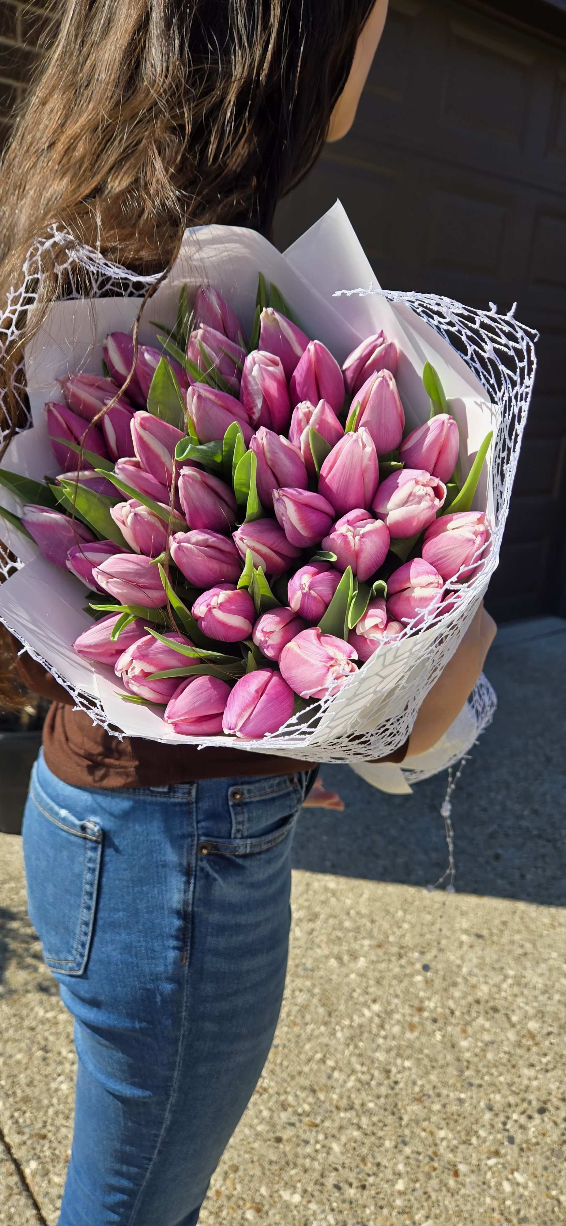 Tulip Embrace  - A stunning bouquet of fresh pink tulips, elegantly wrapped in crisp white paper and delicate mesh. Each bloom is full of vibrant color, symbolizing grace and affection. Perfectly arranged and held under the warm sunlight, this bouquet makes a beautiful statement of love, appreciation, or celebration.  