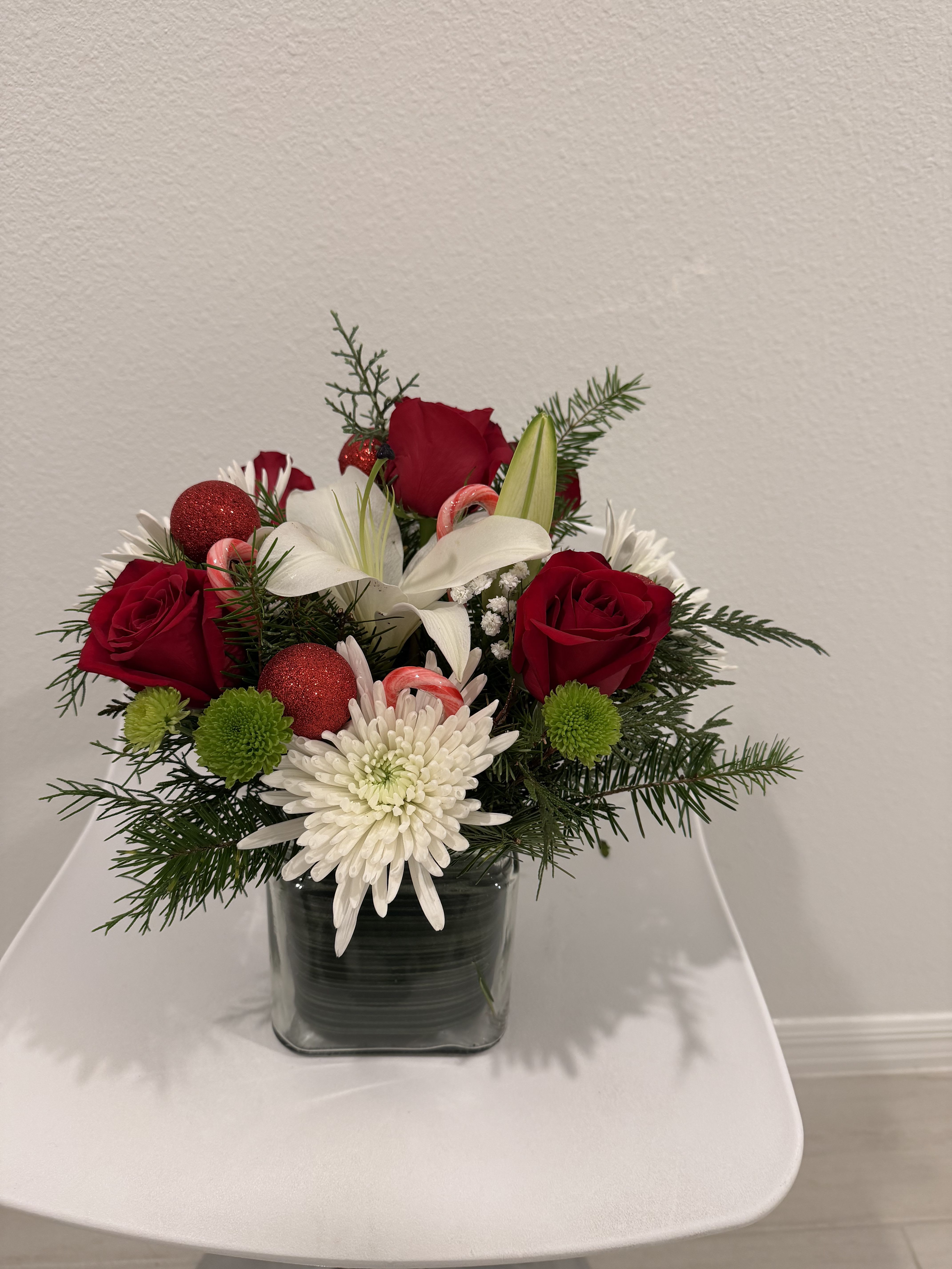 Christmas Dreamlight Bouquet - A joyful holiday arrangement featuring rich red roses, snowy white blooms, bright green accents, and festive evergreens. Adorned with glittering ornaments and candy-cane details, this charming design captures the true spirit of Christmas. Presented in a modern clear glass cube, it brings warmth, beauty, and seasonal bliss to any home or celebration.