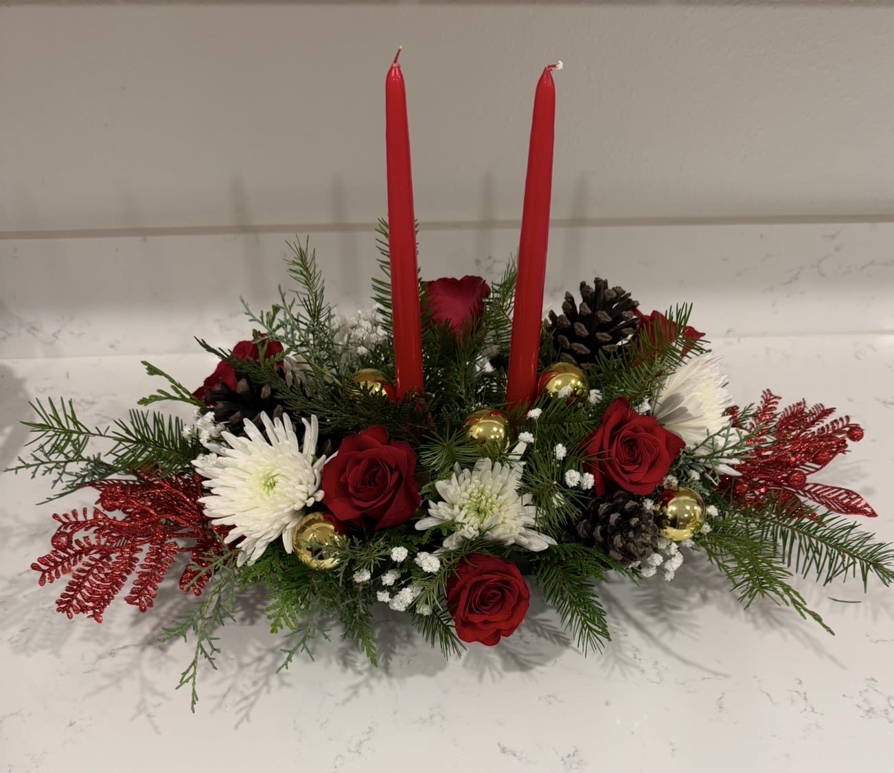 Christmas Candlelight Centerpiece - A refined holiday centerpiece featuring lush evergreens, rich red roses, snowy white mums, and festive accents. Adorned with golden ornaments, pinecones, and sparkling red foliage, this arrangement is beautifully finished with two classic red taper candles. Perfect for adding a warm, glowing touch to your Christmas table or gifting during the season.