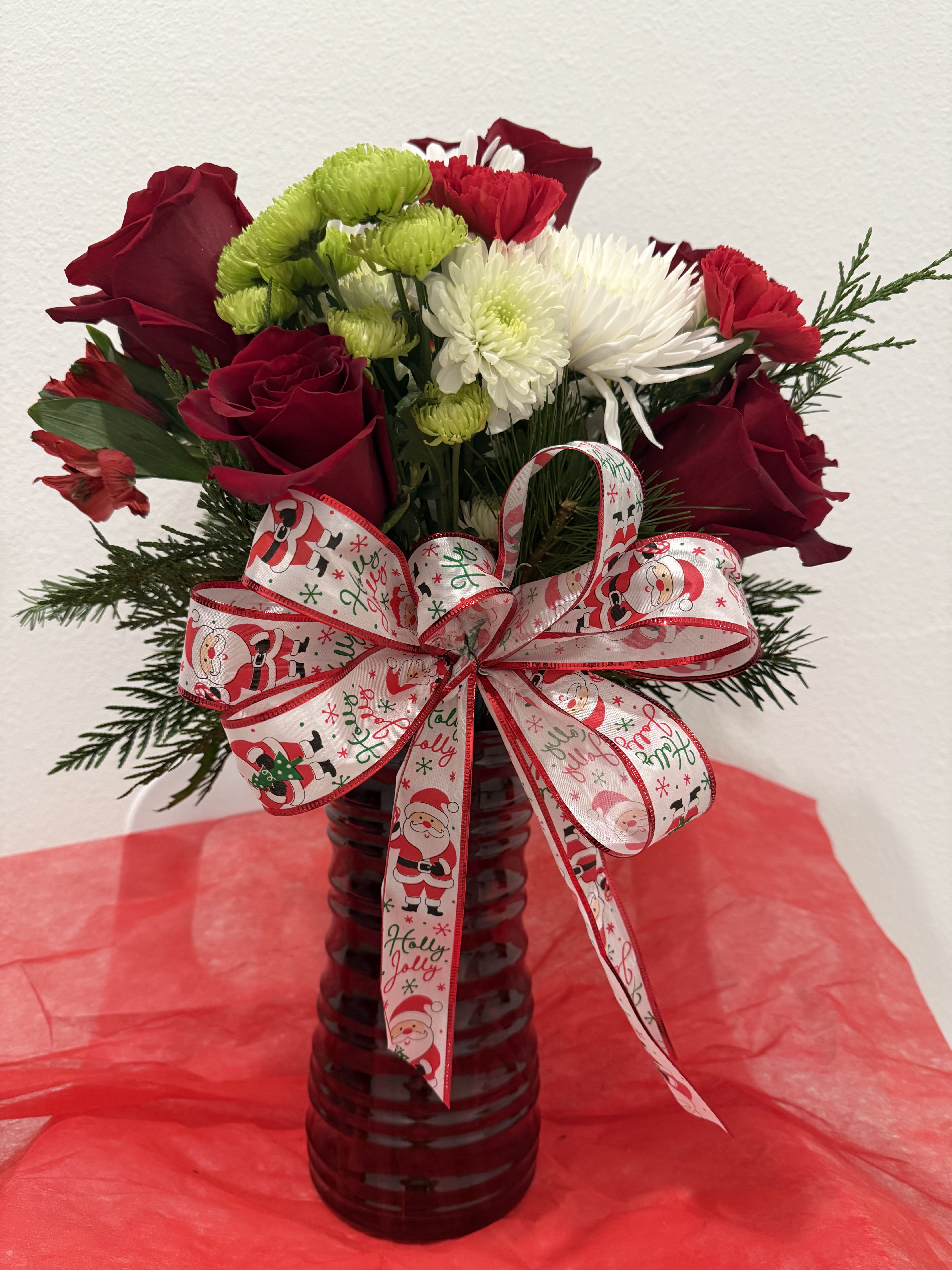 Festive Noel Vase - Celebrate the season with this festive Christmas arrangement featuring rich red roses, snowy white spider mums, cheerful green button chrysanthemums, and seasonal evergreen accents. Designed in a red glass vase and finished with a joyful Santa-themed ribbon, this bouquet brings warmth, color, and holiday cheer to any home or office. A perfect gift to spread Christmas joy and festive spirit.
