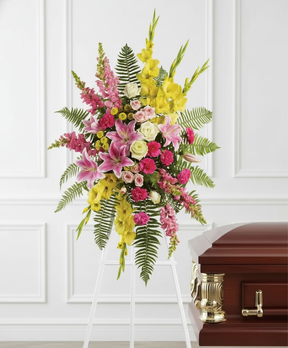 Standing spray of pink lilies, roses, and yellow flowers on a white easel beside a wooden casket.