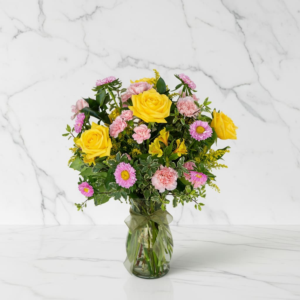 Mixed bouquet of yellow roses and pink flowers in a clear glass vase with a ribbon bow