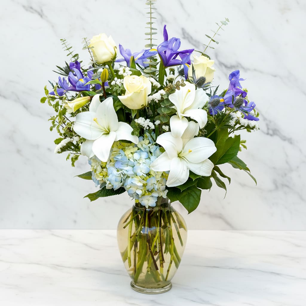White lilies, cream roses, blue hydrangea, and purple flowers in a glass vase