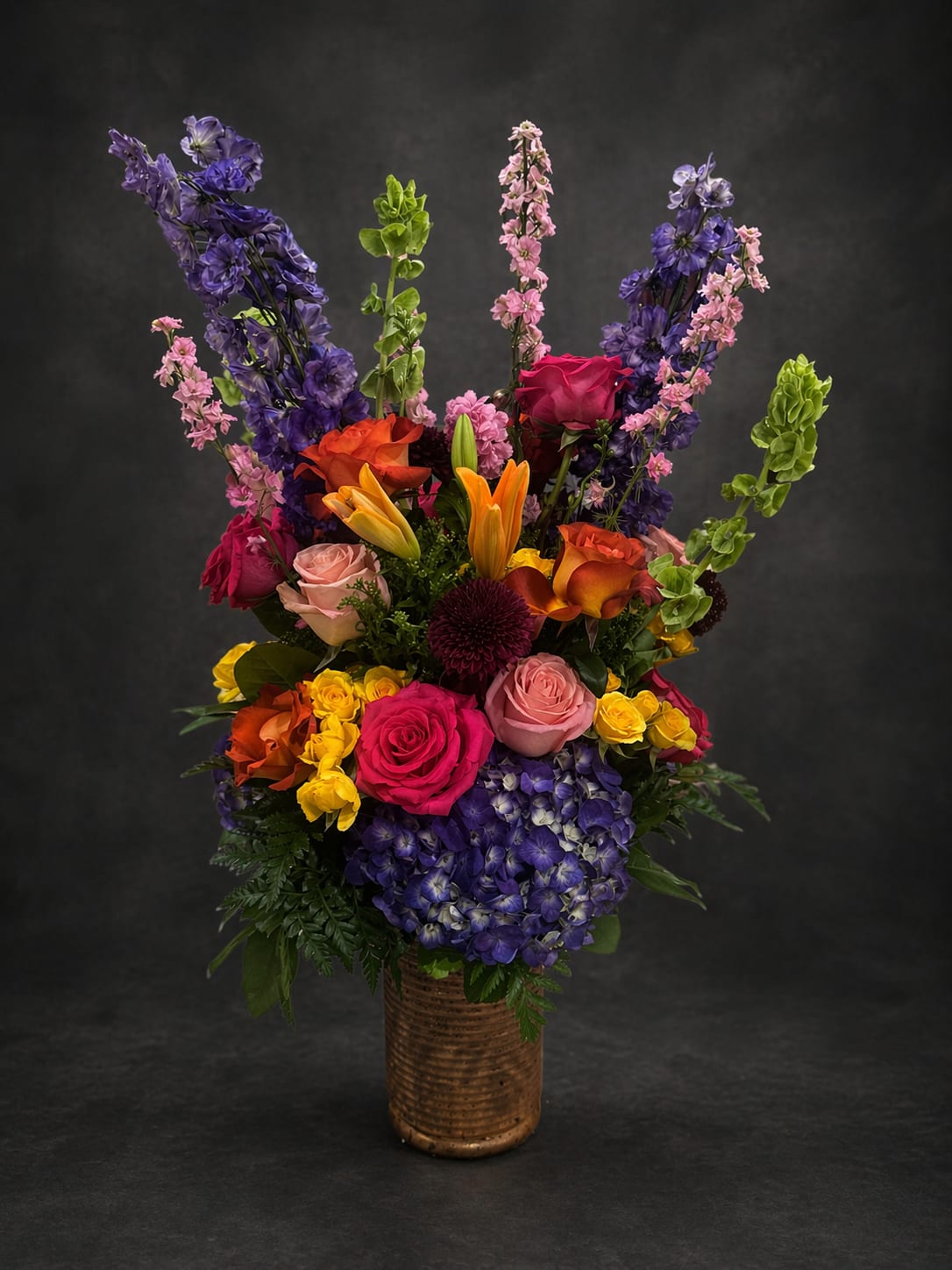 The Love of "Roni" - A tall, jubilant arrangement that reads like a celebration in bloom-perfect as a birthday bouquet or a surprise "just because" gift. The Love of "Roni" combines vertical delphinium spikes with a layered mix of lilies, roses and other seasonal blooms to create a striking display for anniversaries, love and romance, or to let someone know you're thinking of them.  At the base, dense purple hydrangeas form a lush foundation while bright pink, orange, yellow and lavender flowers rise above for vivid contrast and cheerful energy. Rounded center blooms balance the movement of the delphinium spikes, giving the design both height and fullness. Arranged in a ceramic vase, the composition feels both dramatic and refined-an elegant keepsake for sympathy, retirement, thank you, or congratulations moments.  This arrangement is designed to make the recipient feel truly seen-whether you're celebrating a milestone or offering comfort. The tall silhouette adds presence on a mantel or entry table; fresh-cut care is simple-keep water topped up and trim stems every few days for longest life. Order now for a memorable gift that speaks to joy, gratitude, and affection.