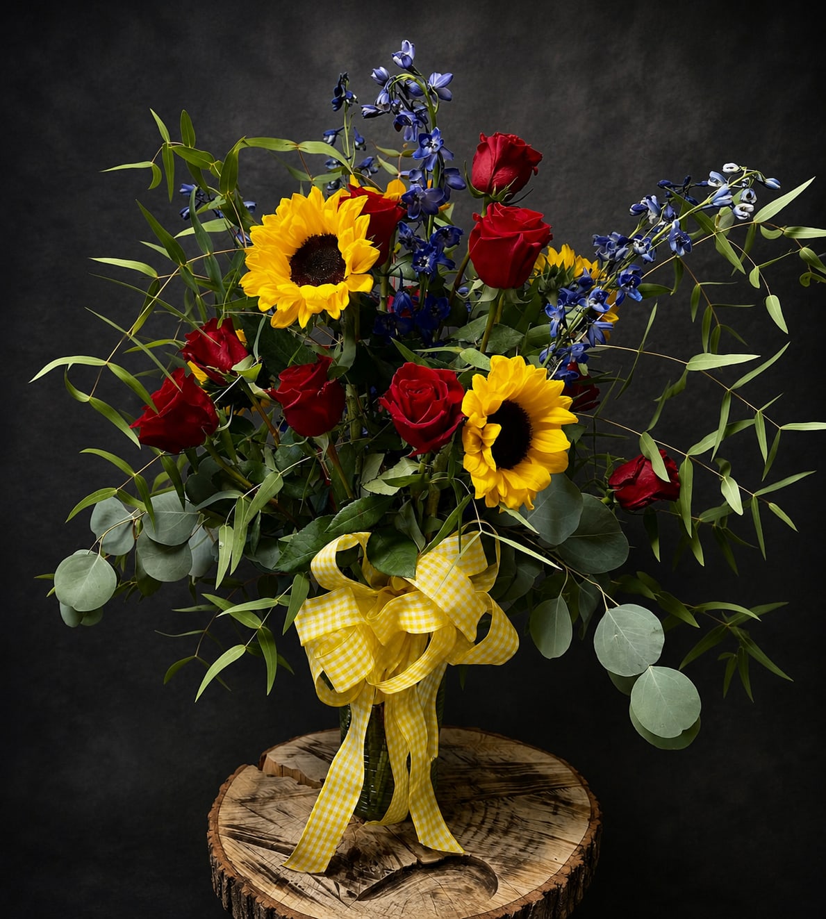 Sunflower, Good Morning - Sunflowers and red roses collide in a bold, high-contrast bouquet that feels like sunshine and celebration in a vase - a perfect birthday, anniversary, or just-because surprise to make the person you're thinking of feel truly seen. The arrangement's warm yellows and passionate reds are playfully balanced by airy blue accent blooms and pale eucalyptus-like foliage, creating a lively, romantic statement that also suits congratulations, get-well wishes, housewarmings, or a cheerful Thanksgiving centerpiece.  Gathered in a clear glass vase and finished with a yellow gingham ribbon, this loose, expansive design features several red roses, bright yellow sunflowers, tall blue accent flowers, and long arching greenery for a full, dramatic silhouette. The texture is both soft and striking: velvety rose petals, bold sunflower faces, and feathery foliage that fans outward. Sizes and vase dimensions match the product images; stems arrive ready to display with simple care-fresh water and a trim of stems every few days keep blooms looking their best.  Whether you need a spirited congratulations for graduation or retirement, a warm Mother's Day or Grandparent's Day greeting, a thoughtful sympathy or thinking-of-you bouquet, or a romantic Valentine or anniversary gesture, this arrangement delivers bright, confident color and the kind of joyful presence that lifts spirits.
