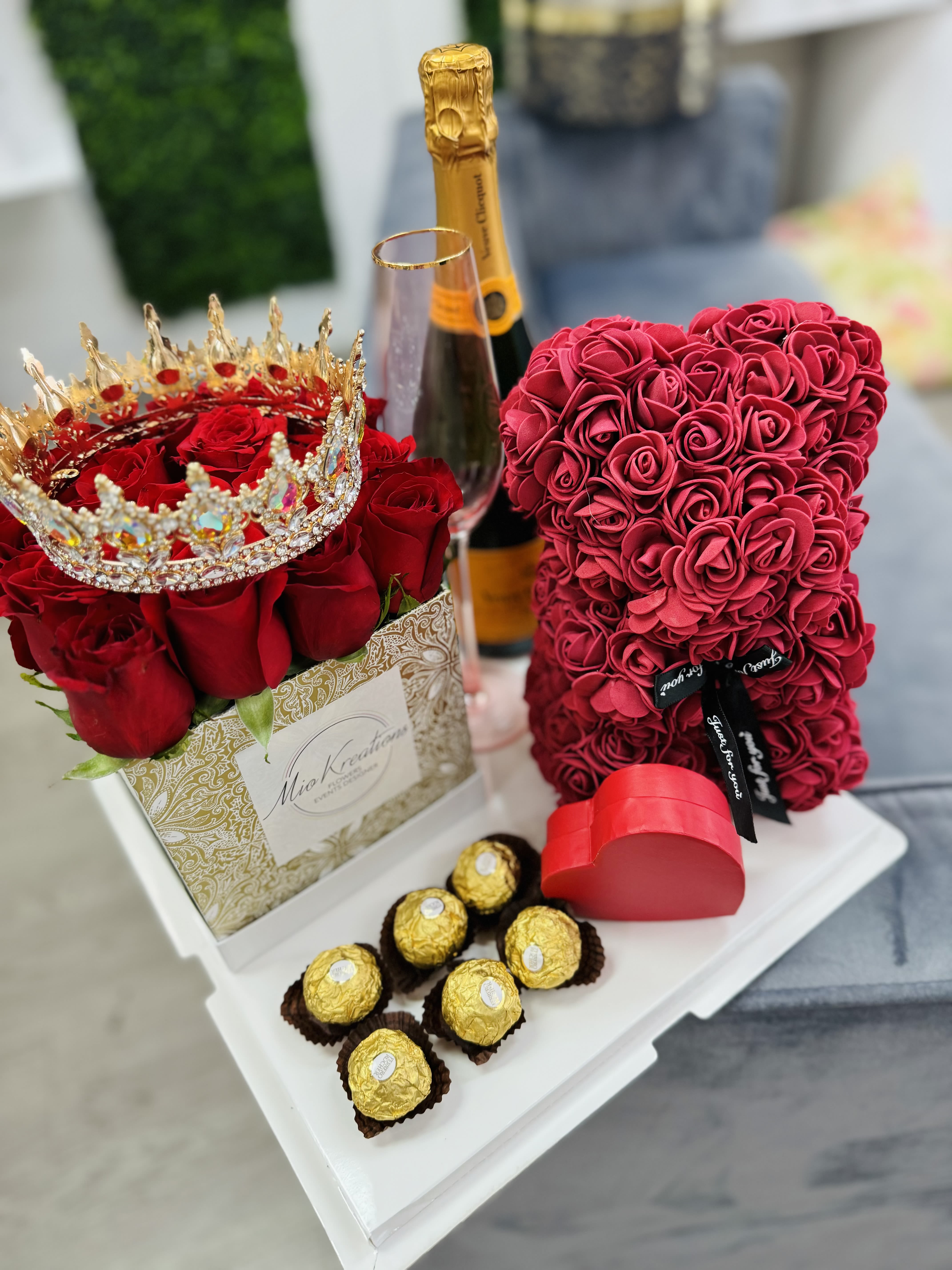 Red rose arrangement with a jeweled crown, champagne bottle, heart box, and chocolates
