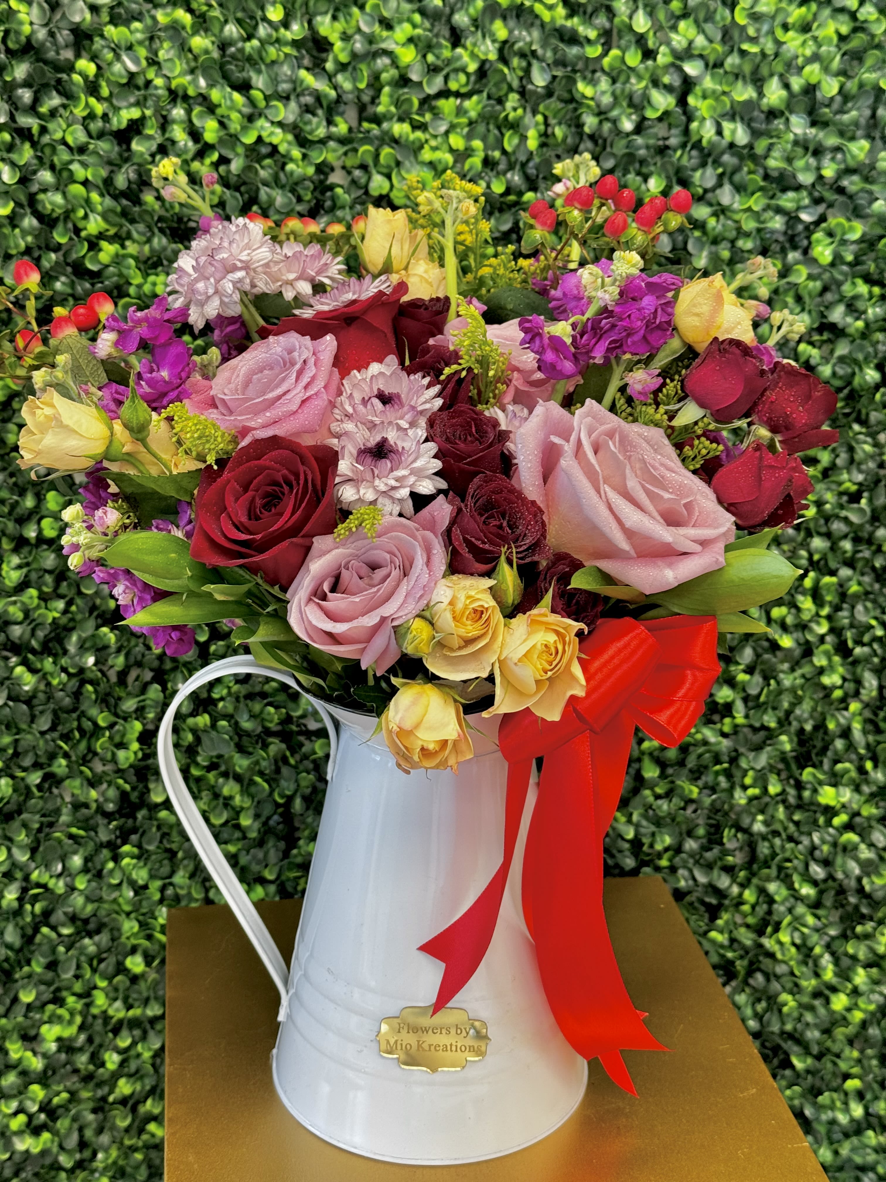 Mixed bouquet of roses and small blooms in a white metal pitcher with a red ribbon