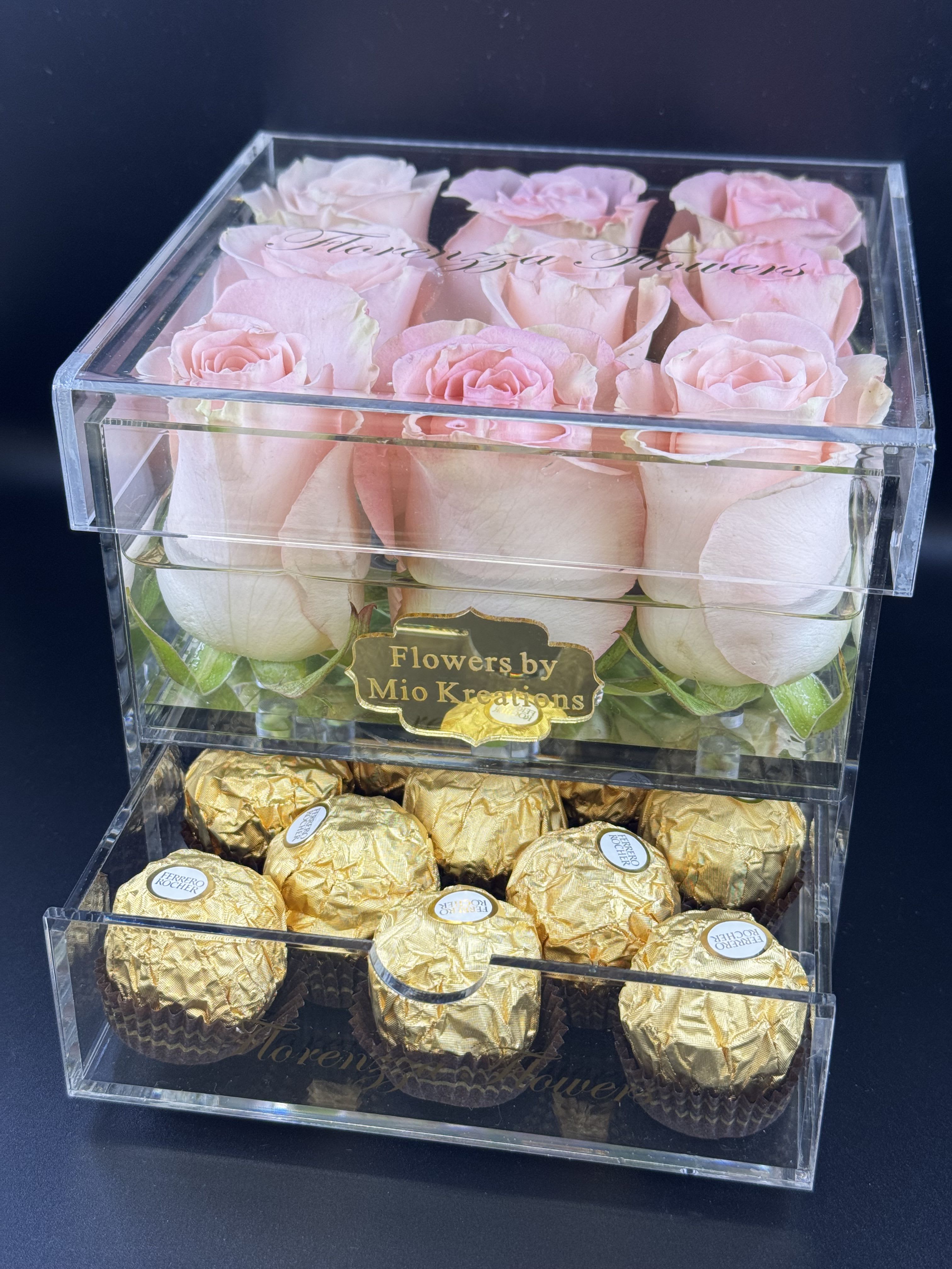 Pink roses in a clear box above gold-wrapped chocolates.