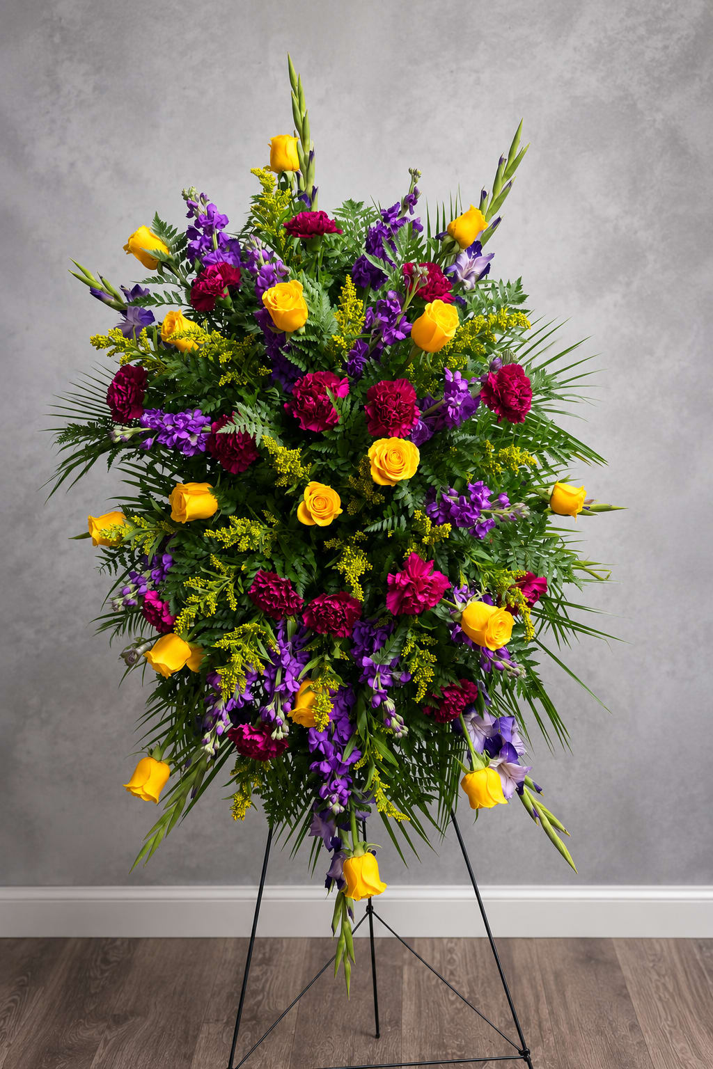 Purple and Yellow easel  - A striking standing spray in yellow, purple, and green designed for sympathy and memorial services-the kind of easel arrangement that offers a bold, reassuring presence at a funeral, celebration of life, or ceremonial tribute. Bright yellow roses draw the eye and lift the mood, while deep purple blooms add depth and solemn richness so the person you're honoring feels remembered and respected.  This large floral arrangement is assembled on a sturdy metal easel and features roses, carnations, stock and seasonal accents for height and texture. Tall spikes and cascading stems create a dramatic vertical silhouette; dense greenery and yellow filler soften the edges for a lush, layered look. The palette of yellow, purple, and green is high-contrast yet harmonious, made to read clearly from a distance in a chapel or reception space.  Made to convey sympathy and heartfelt remembrance, this easel spray is appropriate for family, close friends, or colleagues who deserve a formal tribute. Size and flower varieties may vary slightly with seasonal availability; if substitutions are necessary they will preserve the overall color scheme and style. For delivery to a service or family home, please add service details at checkout to ensure timely placement.