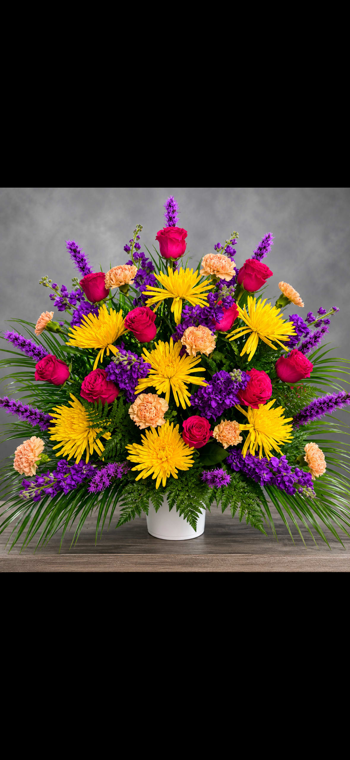 Hot Pink Purple Yellow Funeral Arrangement  - Vivid yellow spider mums and hot pink roses take center stage in this mixed-color funeral arrangement, offering a bright, uplifting tribute that reads as both respectful and celebratory. Designed as a large, full bouquet for sympathy or remembrance, the arrangement's energetic color palette-yellow, pink, purple-brings warmth and comfort to a memorial setting while also suiting celebrations like birthdays or congratulations when a more joyful send-off is desired.  Built in a white ceramic vase, the dense, layered design combines roses, carnations, chrysanthemums, snapdragons and other mixed blooms for a wide, rounded silhouette that feels abundant and thoughtfully composed. Long arching foliage and upright purple spikes add height and graceful movement; yellow carnations and purple accents soften the bold contrasts for a balanced, textured look. The vase provides sturdy support and a clean, elegant base that fits on a mantel, reception table, or altar.  Made to comfort the person you're honoring, this arrangement communicates warmth and remembrance without somberness-the kind of floral expression that helps someone feel seen and celebrated. The large size fills a space with color; for best appearance, keep the vase topped up with fresh water and place away from direct heat or drafts.
