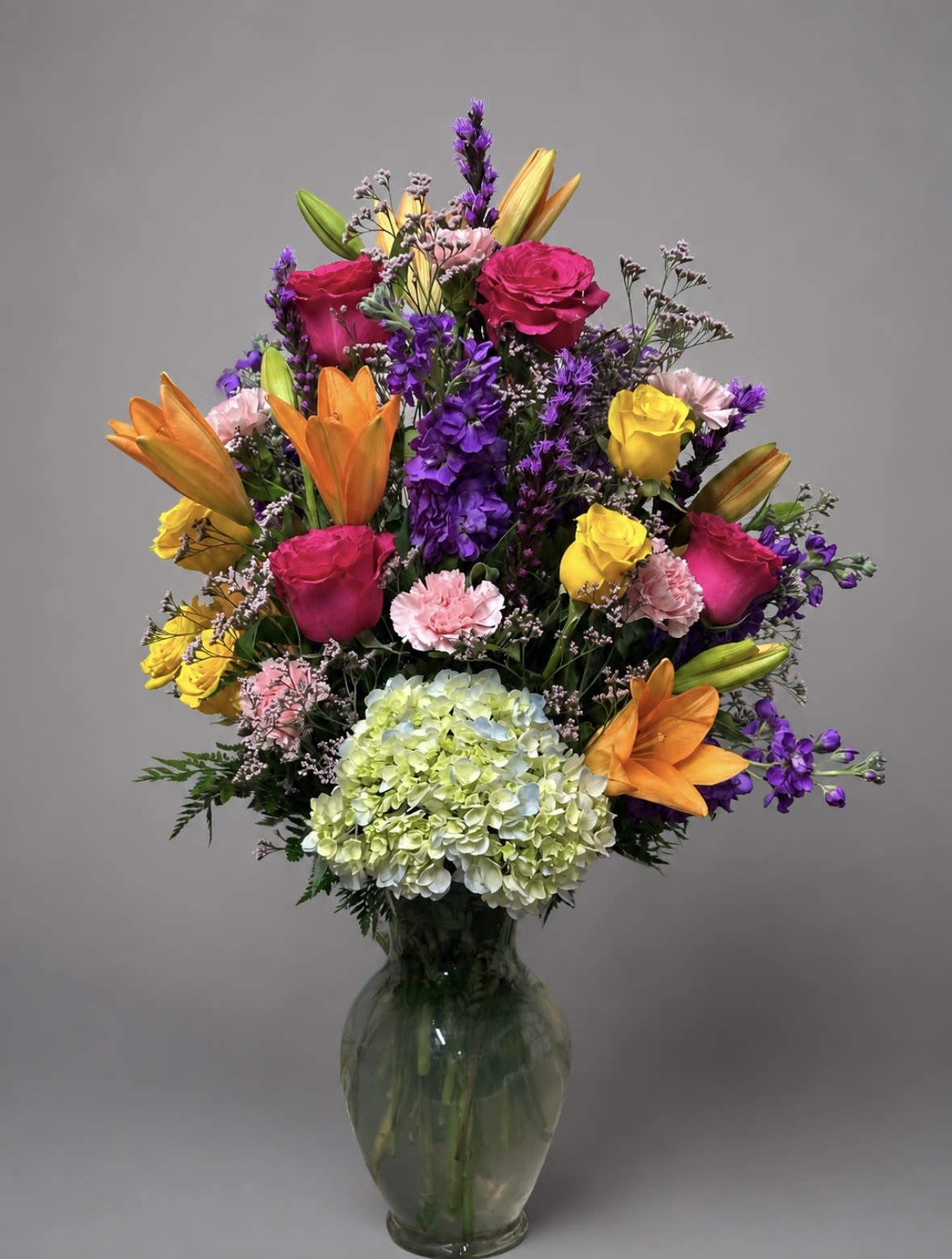 Hot Pink Rose Orange Lily Mix - Vivid hot-pink roses and fiery orange lilies create an immediate burst of color in this mixed-color flower arrangement-perfect as a birthday bouquet, a congratulations gift, or a cheerful thank-you arrangement for someone who lights up a room. The tall, abundant silhouette feels celebratory and luxurious, designed to make the person you're thinking of feel seen and celebrated from the moment it arrives.  This dense, layered design pairs yellow roses, purple stock, pale pink carnations and a large green-to-blue hydrangea for rich contrast and texture, all arranged at varying heights in a clear glass vase for a polished finish. The mix of warm and cool tones gives a lively, festive energy while the glass vase keeps the look refined and display-ready.  Ideal for sending to a colleague, friend, or family member after a milestone or just to brighten their day-this arrangement arrives full and ready to enjoy. Standard care: keep in fresh water, out of direct sun, and trim stems occasionally to extend vase life.