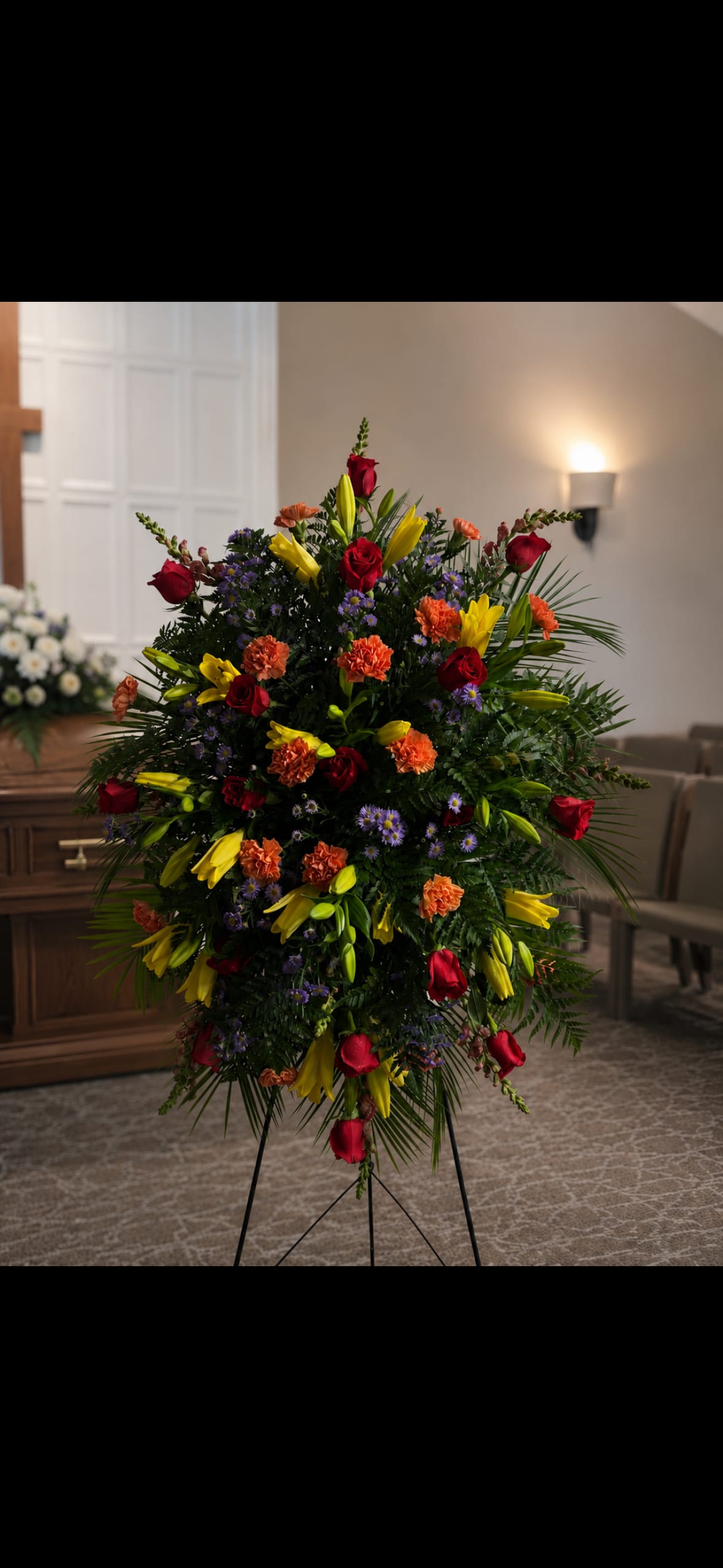Red Orange and Yellow Easel - A bold, ceremonial standing spray that honors a life with vivid warmth and presence-this red, orange and yellow easel arrangement is designed for sympathy and memorial services or any time you need a formal floral tribute. The mix of red roses, yellow lilies, orange carnations, and purple aster accents reads like a heartfelt salute: the colors speak of love, remembrance, and comfort while the easel gives it dignified visibility at a service or graveside.  Dense and rounded in silhouette, the spray features clusters of velvety red roses and ruffled orange carnations framed by bright yellow lilies and delicate purple asters set against deep green foliage. The arrangement is mounted on a sturdy standing easel to create a striking vertical display that's meant to be seen from a distance and placed near caskets, memorials, or ceremony sites. For ordering, choose this piece when you want a powerful, color-rich tribute that ensures the person you're honoring feels remembered and celebrated.