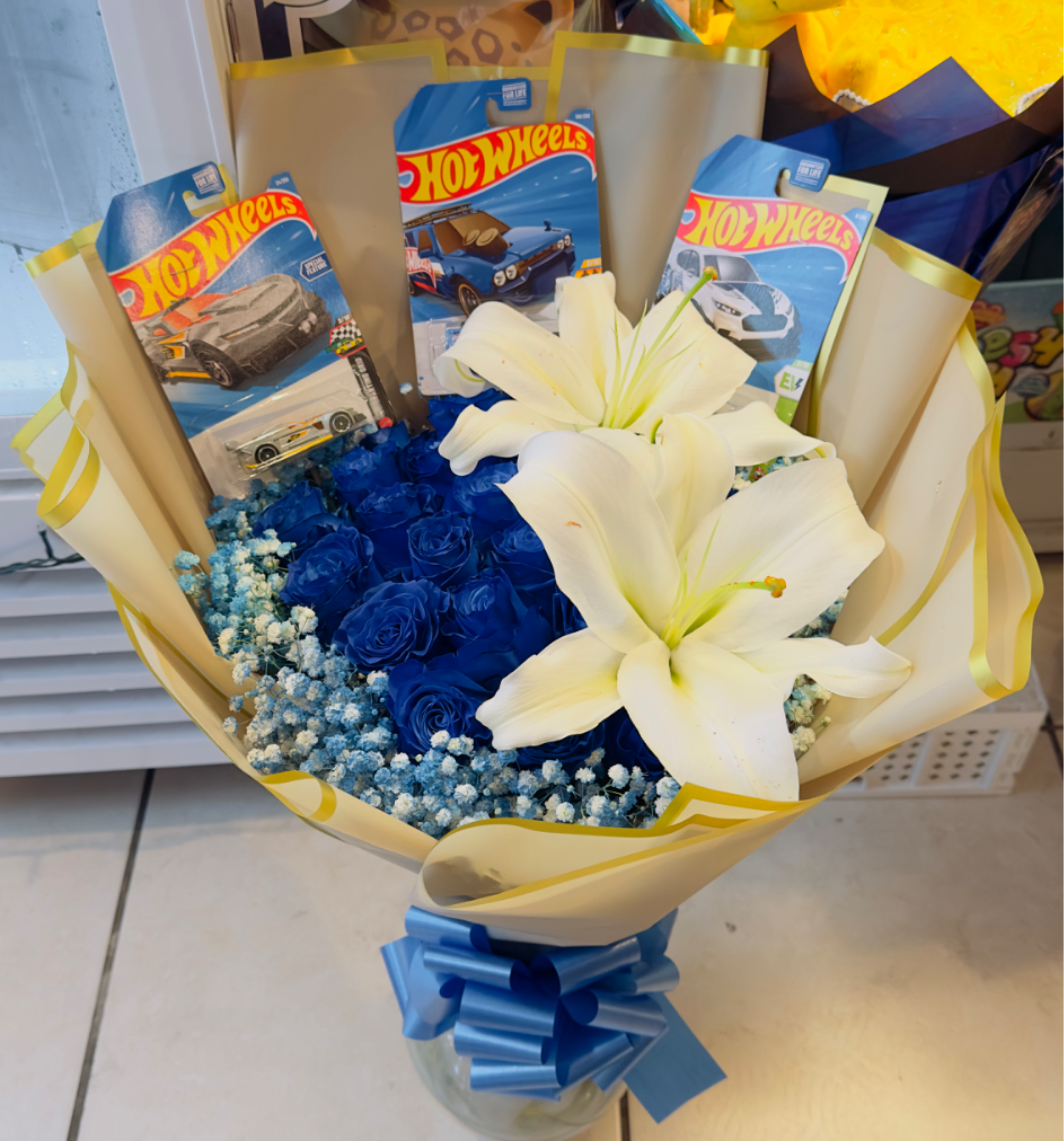 Hot wells  - Start with a show-stopping cluster of vivid blue roses and two large white lilies that create eye-catching contrast-the perfect birthday bouquet or anniversary surprise for someone who loves bold color and playful details. Designed as a gift-forward arrangement, the mix of saturated blues, crisp ivory lilies, and light blue filler flowers reads joyful and energetic, making it ideal for birthdays, graduations, "just because" moments, or sending romantic congratulations with a fun twist.  This arrangement is presented in a glass vase, wrapped in cream-and-gold decorative paper with yellow edging and tied with a wide blue ribbon for a polished, gift-ready look. Several Hot Wheels toy car packages are tucked into the bouquet, turning the flowers into a themed keepsake that's especially suited to kids, car fans, dads on Father's Day, or anyone who appreciates a nostalgic, boyish flair. Size and handling instructions are provided at checkout; for best enjoyment, refresh the vase water every couple of days and trim stems slightly to keep blooms vibrant.
