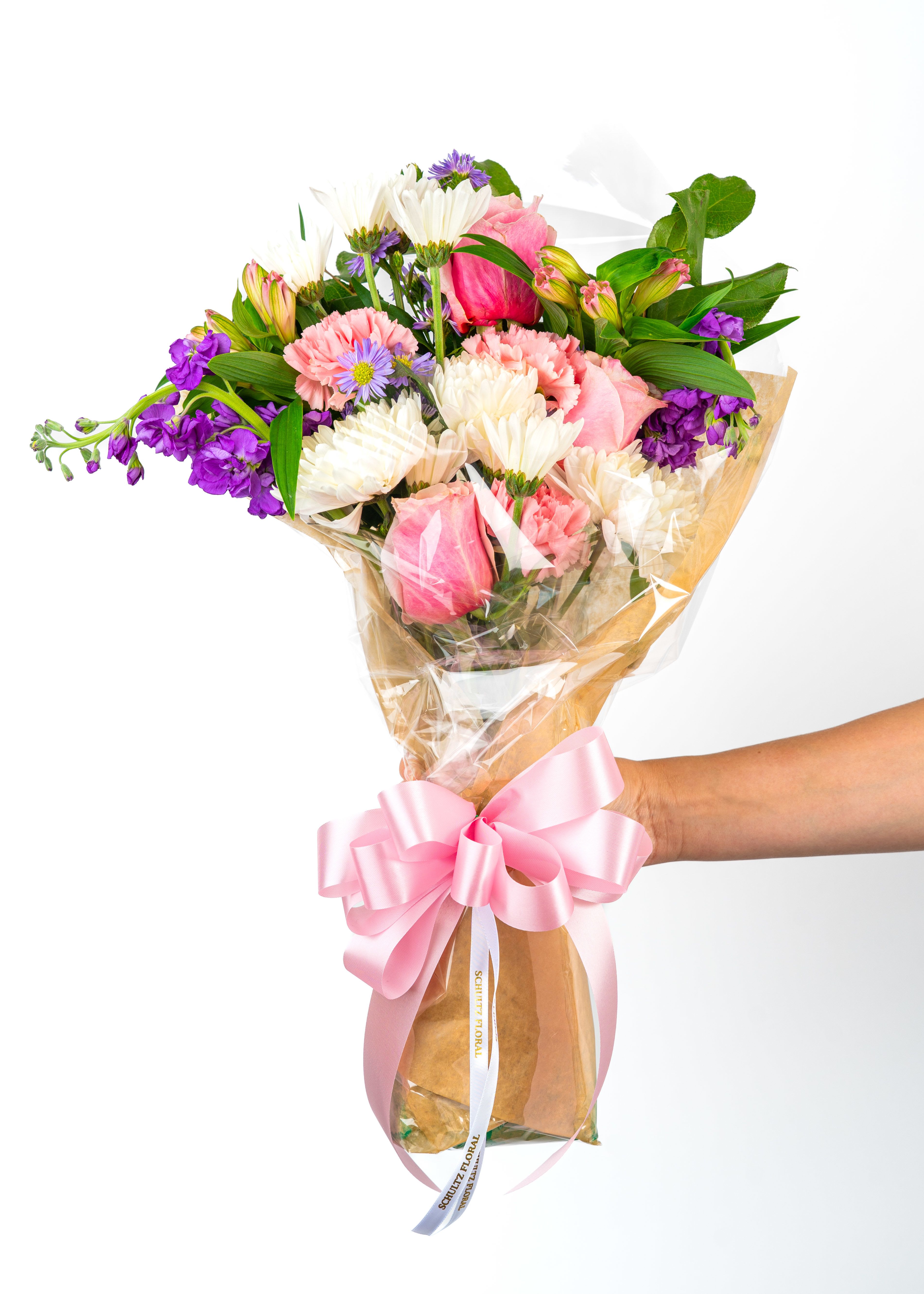Handheld bouquet of pink, white, and purple mixed flowers wrapped in cellophane with a large pink bow