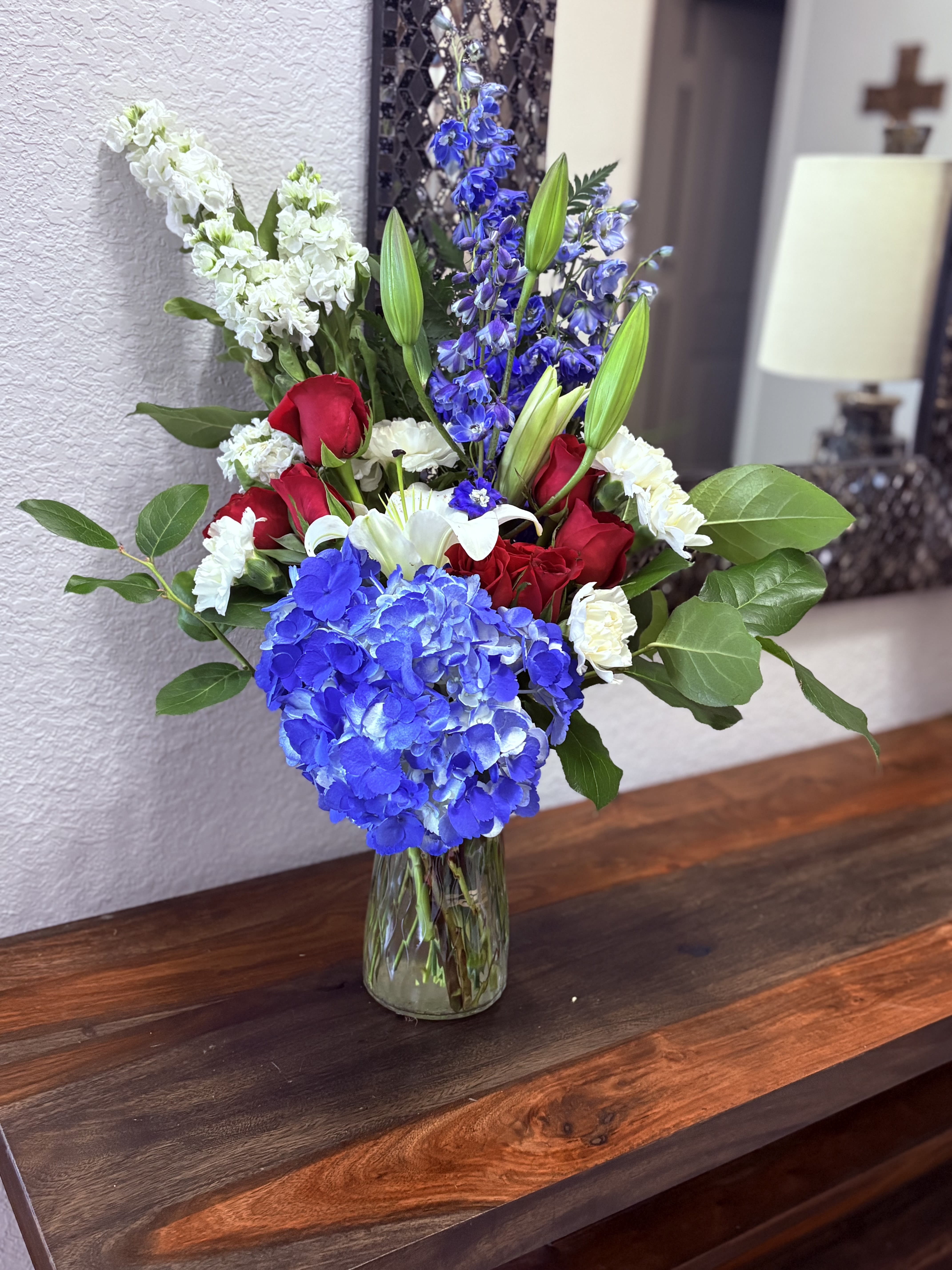 Dance with me bouquet - A bold cluster of blue hydrangea meets classic red roses and white lilies in a formal, celebratory bouquet-perfect as a birthday bouquet, anniversary arrangement, or a thoughtful sympathy display. The "Dance with Me" bouquet's high-contrast palette of saturated blue, crimson, and pure white creates an immediate visual impact that will make the person you're thinking of feel honored and truly seen.  The arrangement features a large blue hydrangea at the front, framed by red roses and elegant white lilies, with tall blue stock rising behind for height and movement. Broad green foliage adds fullness and structure, all gathered in a clear glass vase that showcases the clean, polished composition. Sized to sit proudly on a dining table or mantel, it's a timeless mix of texture and color that reads both festive and refined.  Ideal for celebrating milestones or expressing heartfelt sympathy, this bouquet sends a clear message of respect and affection. Vase-ready and arranged by hand, it arrives ready to display-lightly mist the blooms and keep them in a cool spot away from direct sunlight to extend their freshness.