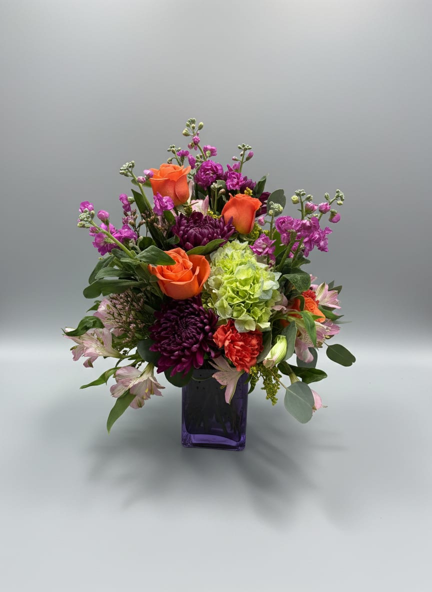 Sweet bloom - Bright bursts of orange roses meet vivid purple blooms and a lime-green hydrangea cluster to create a joyful, celebratory flower arrangement perfect for birthdays, congratulations, or a cheerful everyday gift. This flower arrangement for spring gifting places the recipient at the center-she'll feel seen and celebrated by the bold contrast of warm orange and cool purple, softened by pale pink accents.  The design features roses, hydrangeas, carnations, alstroemeria, chrysanthemums, stock and other mixed seasonal blooms arranged in a rounded, garden-style composition. A deep purple glass vase echoes the purple flowers and gives the piece a modern, polished base while the airy branching accents and layered focal flowers add texture and movement. Sized to read full and abundant, it's an ideal pick for a colorful home display or as a vibrant thank-you or birthday surprise-simply refresh the water every few days and trim stems for longer-lasting blooms.