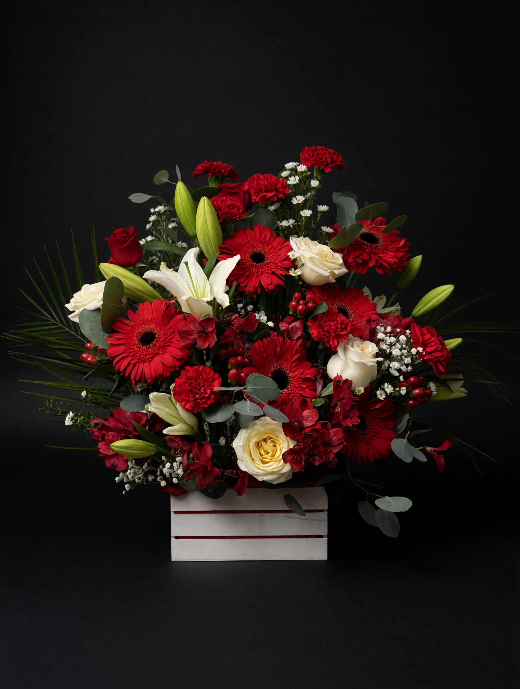 Red sympathy  - A bold red-and-white crate arrangement that reads as both festive and deeply heartfelt-perfect as a romantic birthday bouquet, a Christmas centerpiece, or a respectful sympathy tribute. The striking mix of red gerbera daisies, red carnations and red roses against clusters of white roses and white lilies creates a dramatic, classic contrast that makes the recipient feel genuinely seen and honored.  Tightly layered in a white wooden crate, the composition rises above the container with tall lily buds and airy filler blooms for height while eucalyptus and berry accents soften the silhouette with gray-green texture. The palette of deep crimson, pure white and fresh greens gives a polished, formal look; the arrangement's abundant shape and dense flower coverage deliver a lush, high-impact presence for mantels, tabletops, or beside a casket.  Ideal for someone you want to celebrate, console, or impress-this piece says love, comfort, and timeless elegance. Sizes are designed to create a broad, abundant display; water and remove any wilted blooms as needed to keep lilies and roses looking fresh. Order early for holiday or memorial delivery to ensure this statement arrangement arrives at its peak.