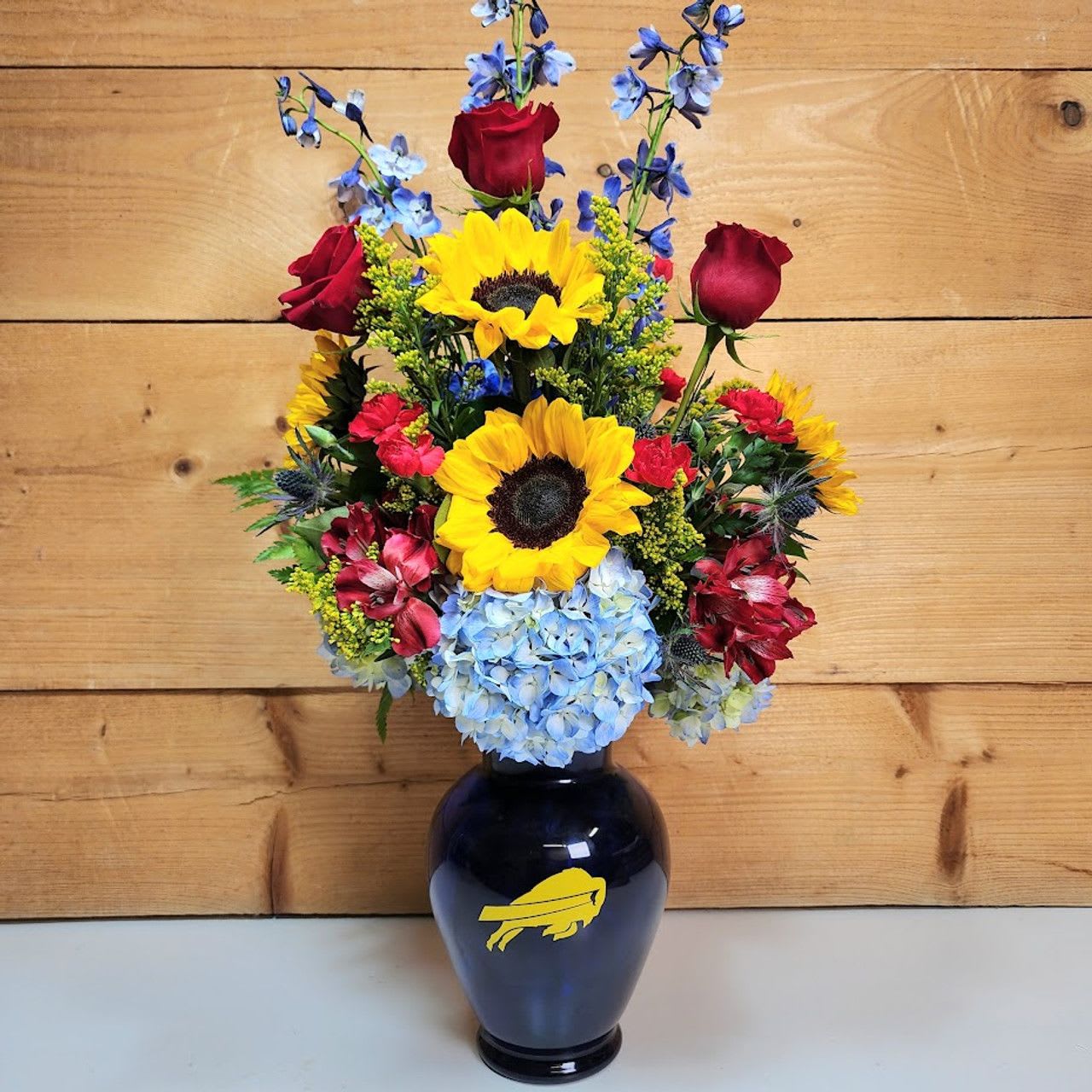 Bouquet of sunflowers, red roses, blue hydrangea, and blue delphinium in a dark vase