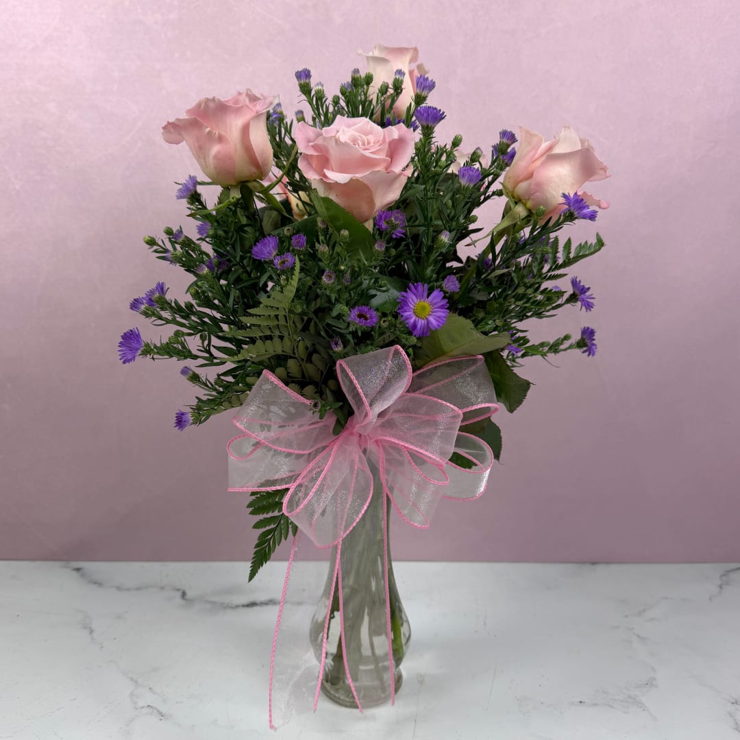 Pink roses and purple daisies in a clear glass vase with a pink ribbon bow.