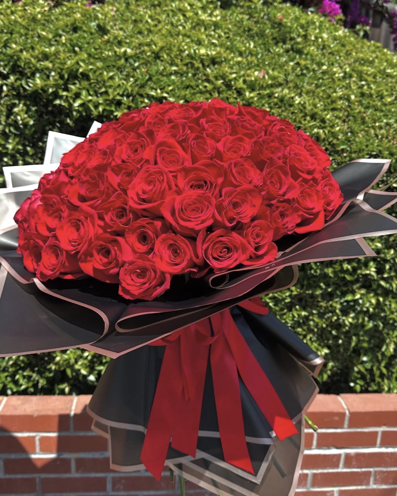 100 Ways to Say “I Love You” - Make a grand romantic statement with this breathtaking hand-tied bouquet of 100 luxurious red roses. Each rose is perfectly arranged to create a lush, full bloom that speaks volumes of love, passion, and devotion. Wrapped in elegant black designer paper with a striking red satin bow, this bouquet is a timeless expression of affection—because sometimes, one hundred roses say it better than words ever could.